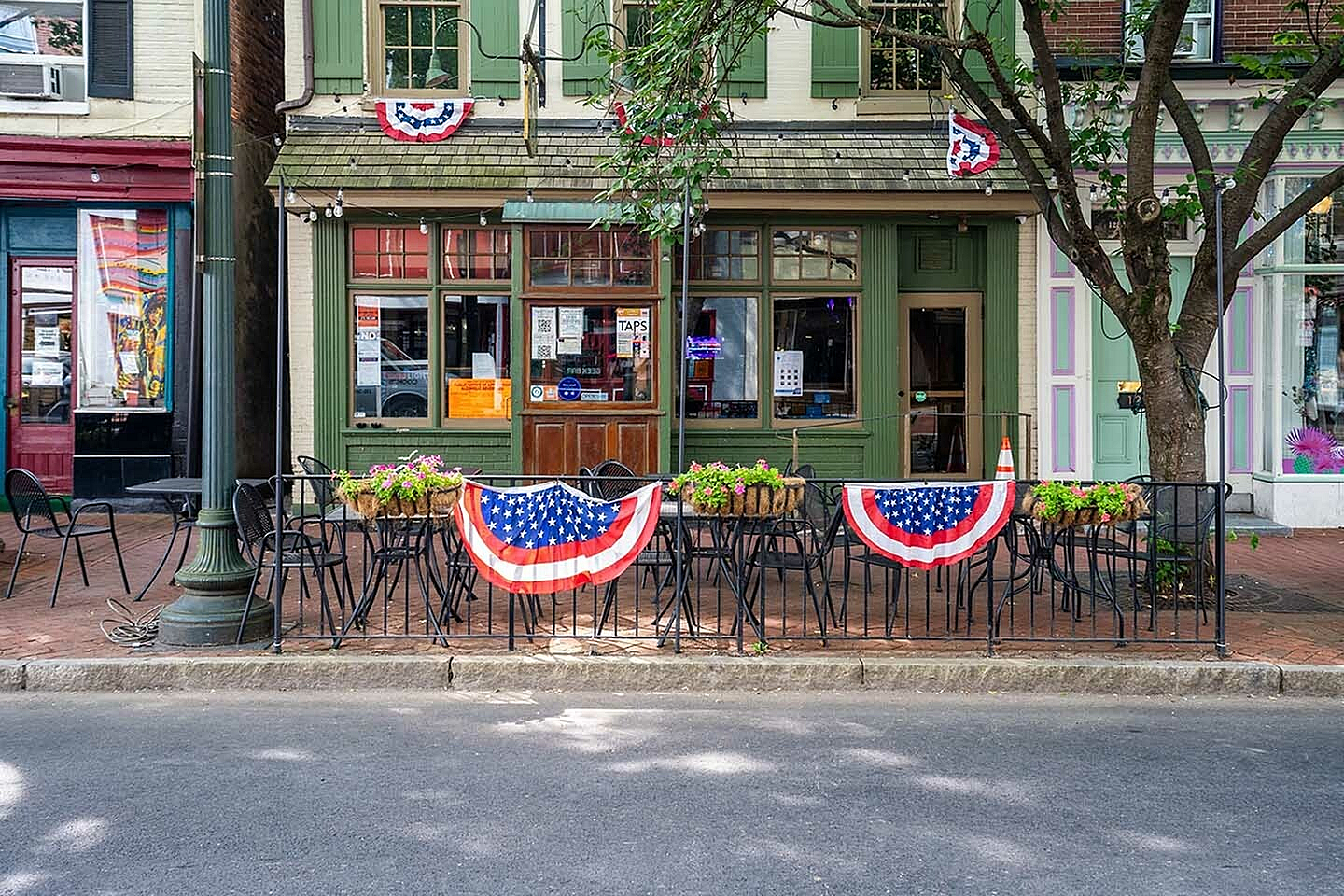 Charming outdoor café adorned with American flags, featuring cozy seating and lush planters on a vibrant city street.