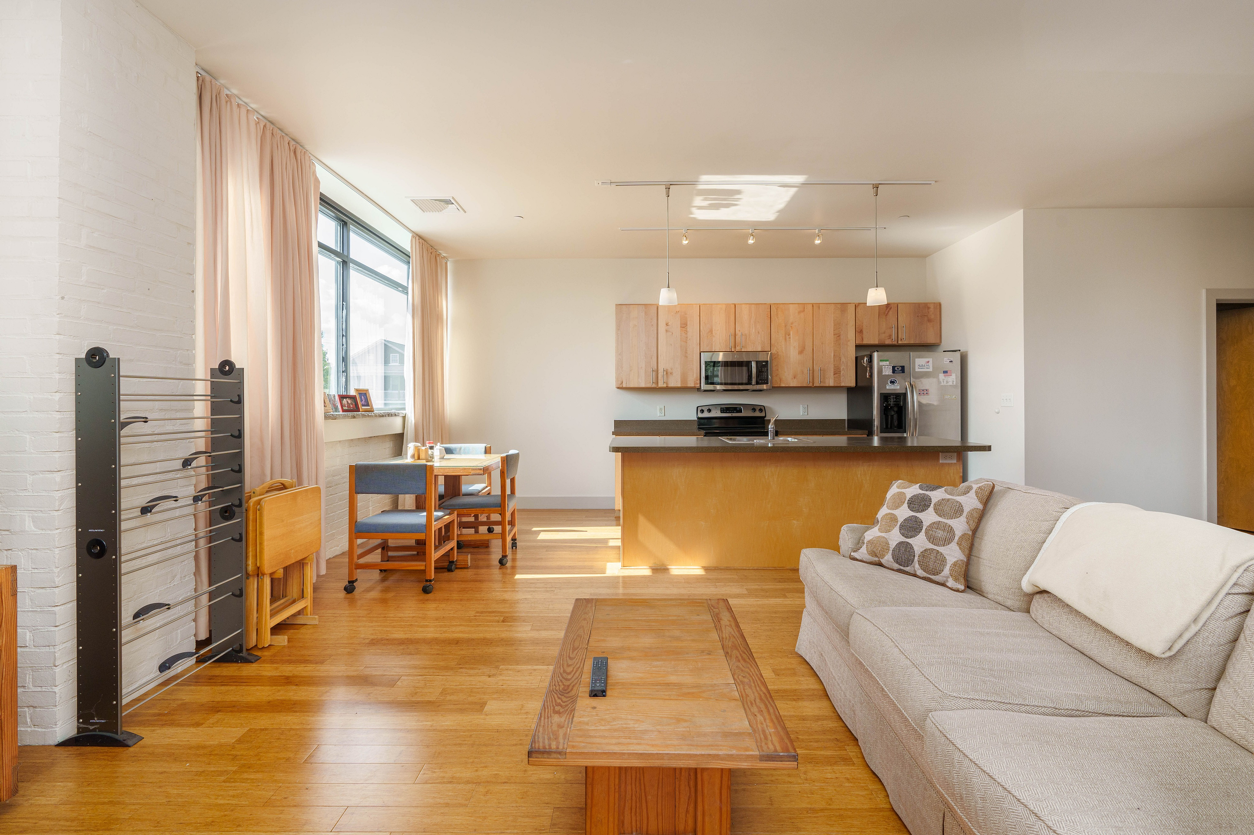 Bright and modern open-plan living area featuring a minimalist kitchen with wooden cabinetry, cozy lounge with a comfortable beige sofa, and a sunlit dining space near large windows.