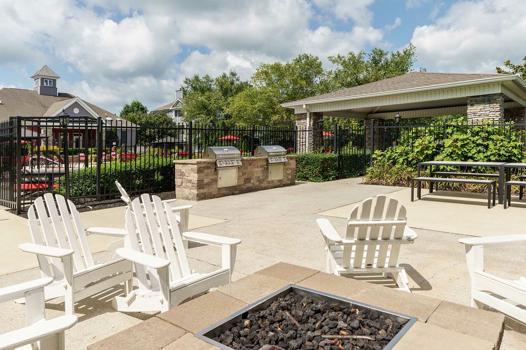Charming outdoor patio with cozy white Adirondack chairs around a modern fire pit, adjacent to a built-in grill area and surrounded by lush greenery.