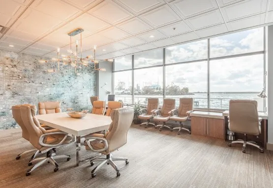 Elegant office space with a stylish meeting table, comfortable chairs, and floor-to-ceiling windows overlooking a waterfront view.