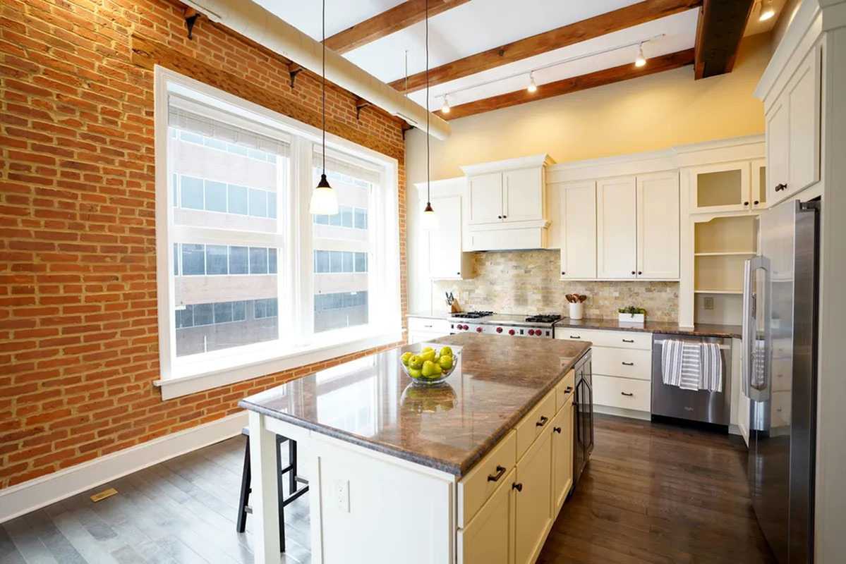 Charming kitchen with exposed brick walls, elegant white cabinets, and a large island. Features natural light from expansive windows and stylish pendant lights.