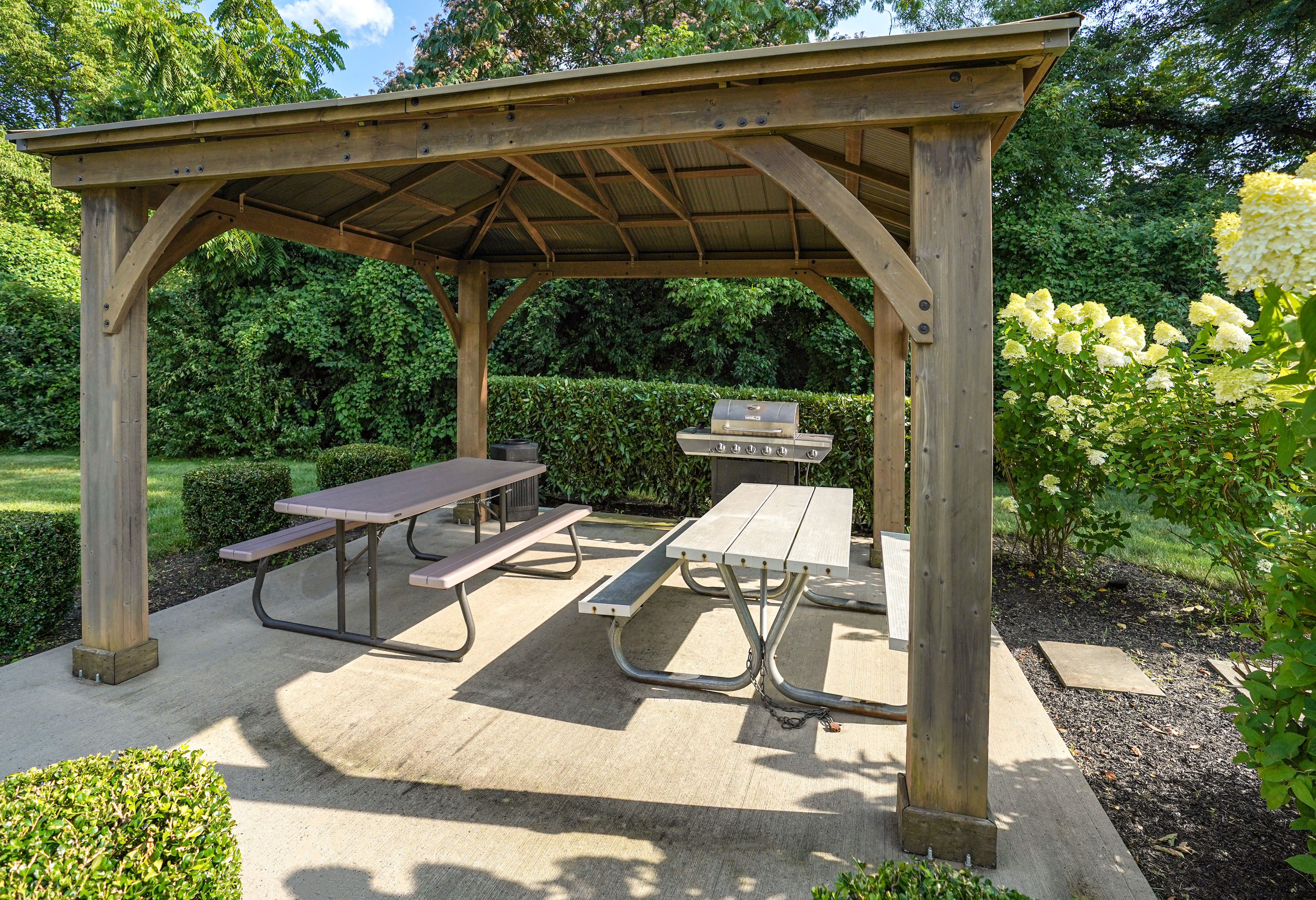 Charming outdoor pavilion with picnic tables and a grill, nestled in lush greenery and blooming flowers, ideal for gatherings and relaxation.