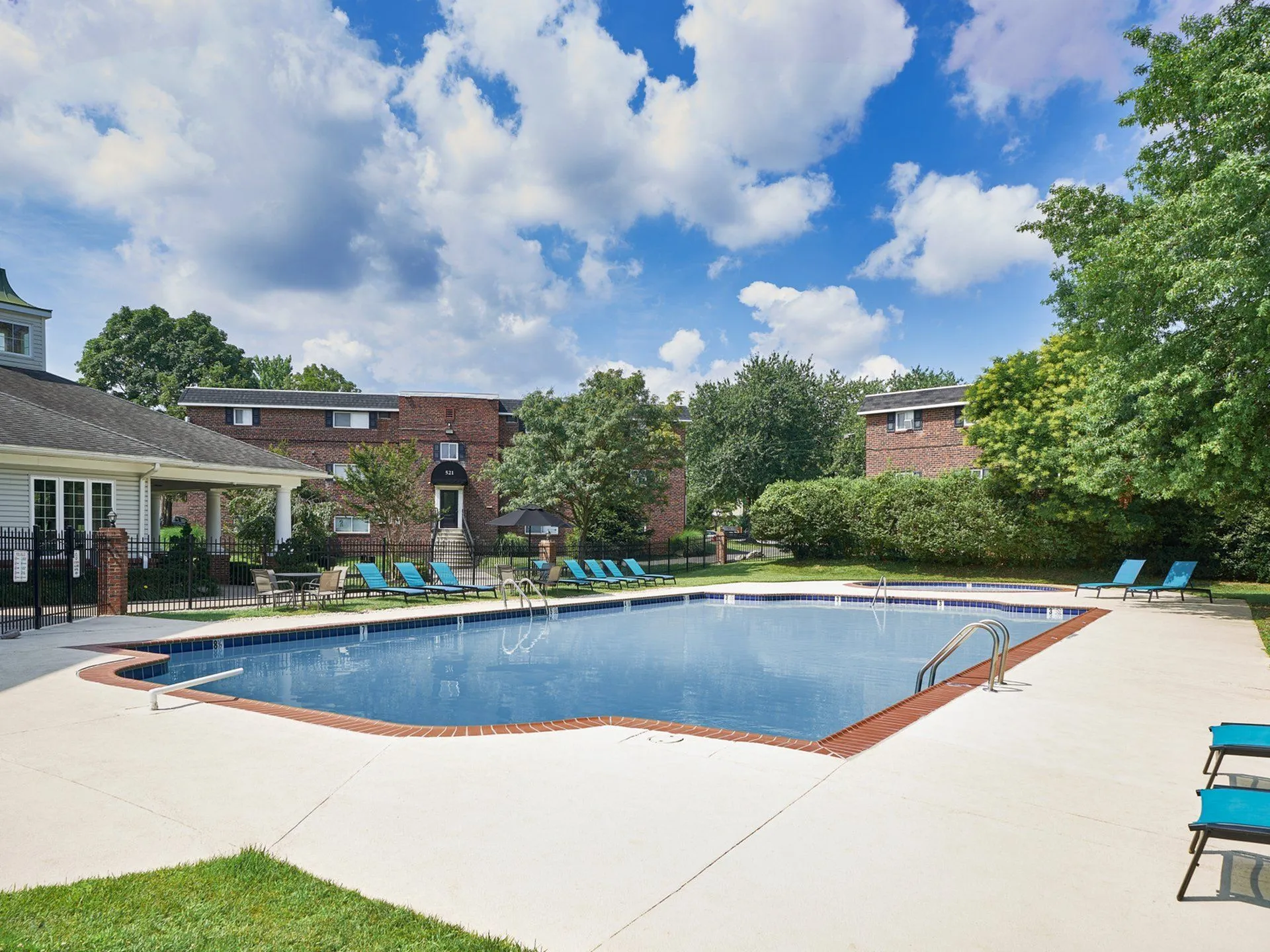 Inviting outdoor pool area with sun loungers, surrounded by lush trees and charming brick buildings.