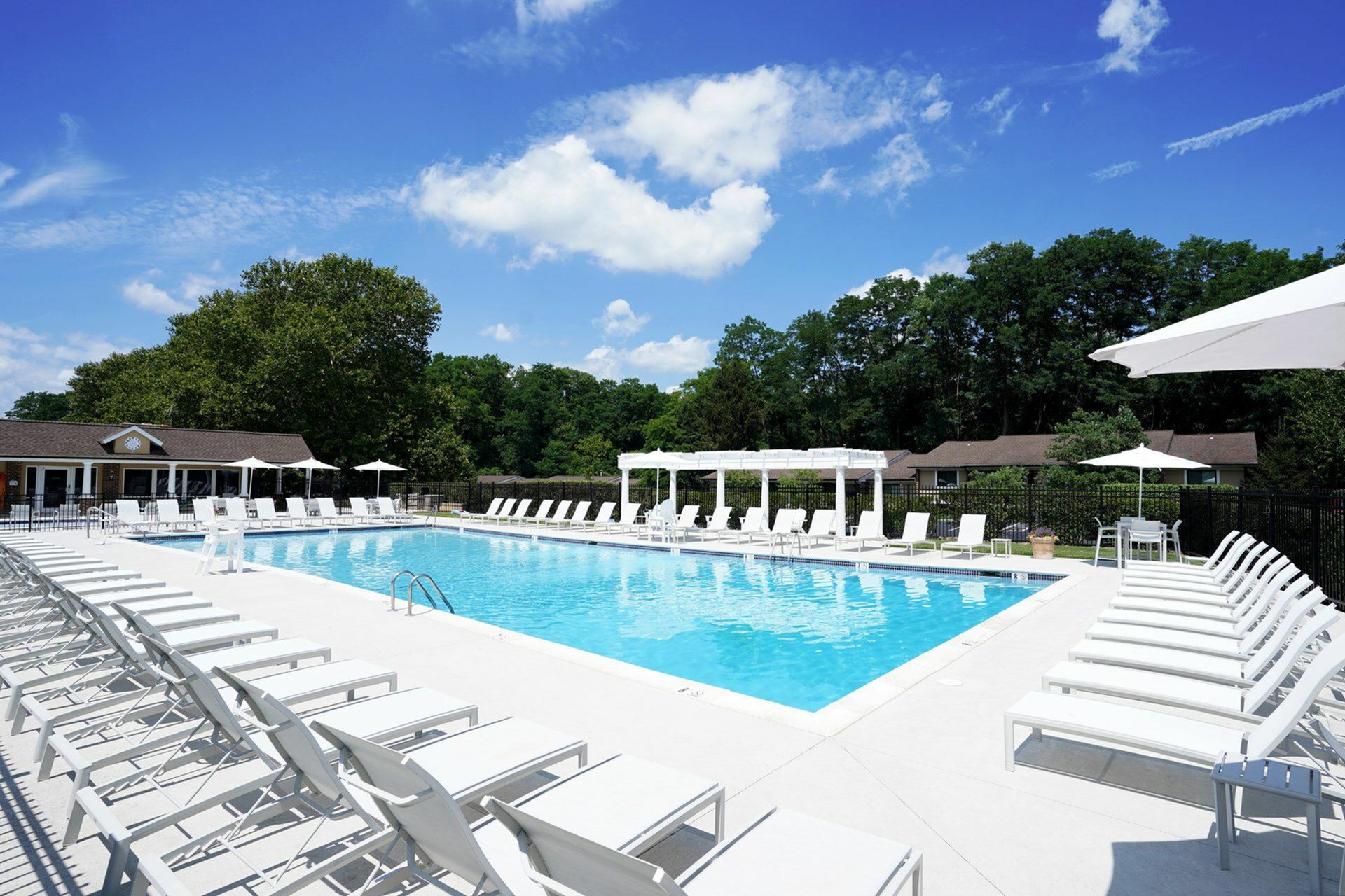 Luxurious outdoor swimming pool with modern white lounge chairs and umbrellas, surrounded by lush greenery and a vibrant blue sky.