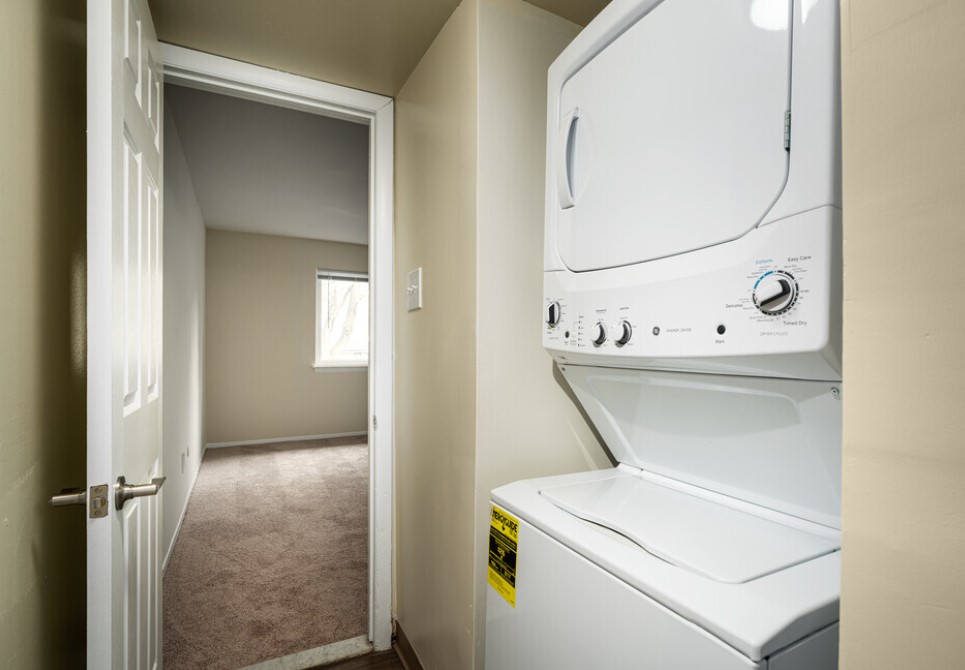 Contemporary apartment hallway featuring in-unit stackable washer and dryer next to a cozy, carpeted room with a bright window.