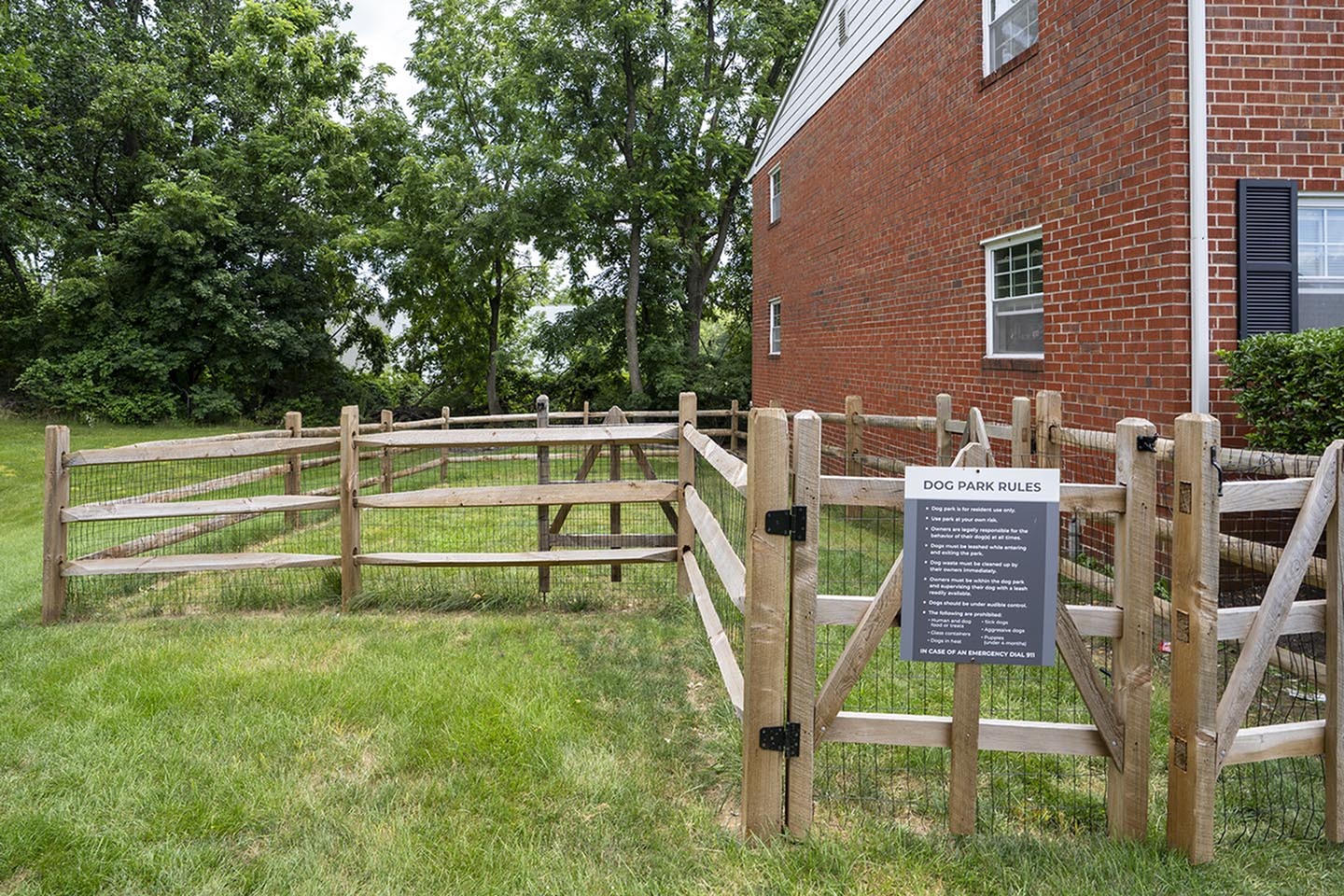 Inviting dog park with wooden fencing beside a charming brick apartment, surrounded by lush greenery.