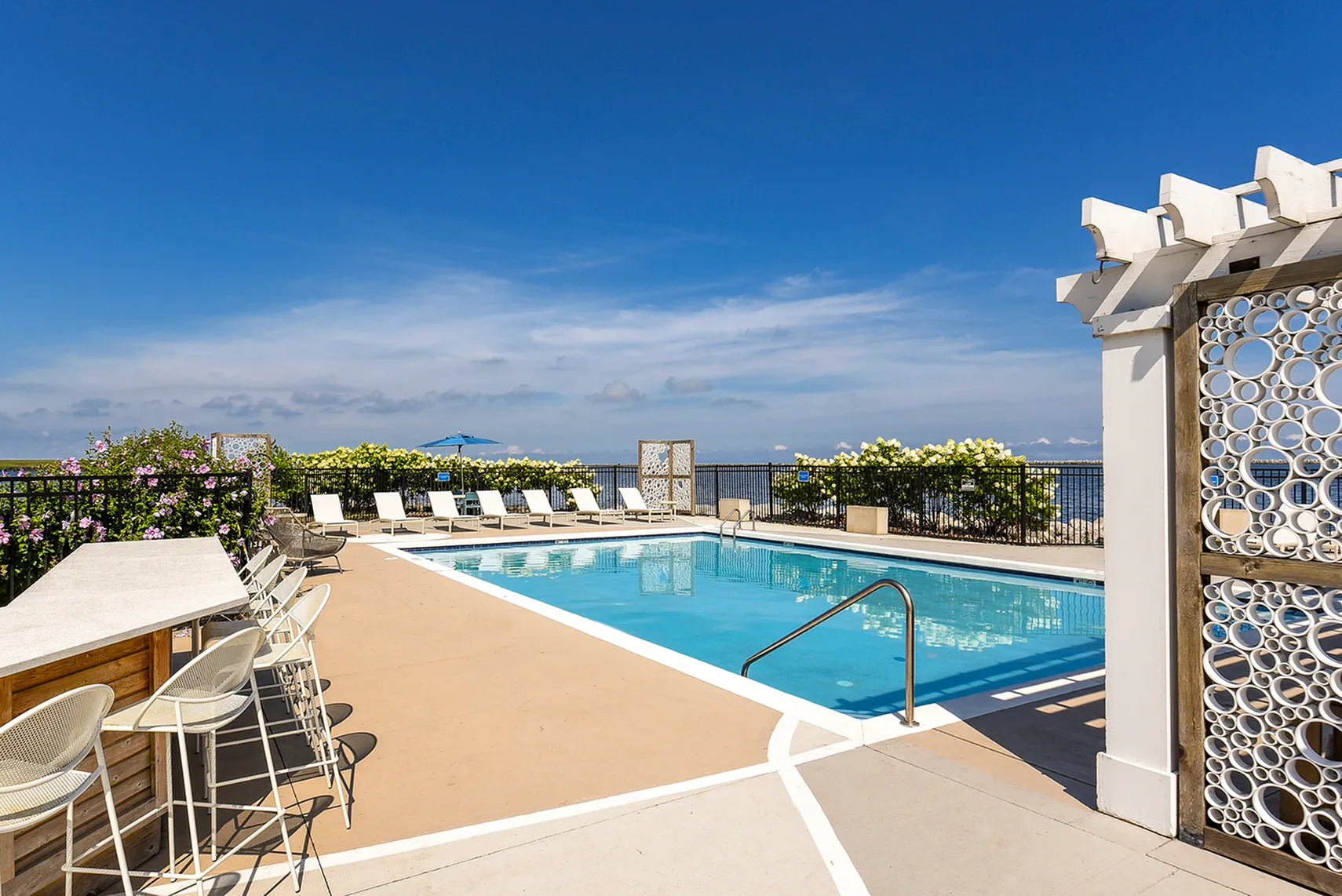 Luxurious rooftop pool area with stylish lounge chairs, scenic ocean view, and vibrant landscaping against a clear blue sky.