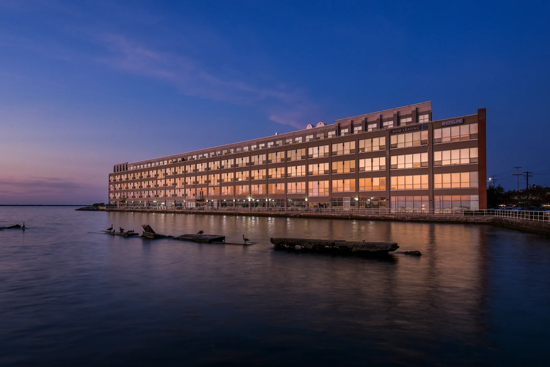 Elegant waterfront building with modern glass fa&ccedil;ade reflecting vibrant sunset hues, enhancing upscale living by the water.