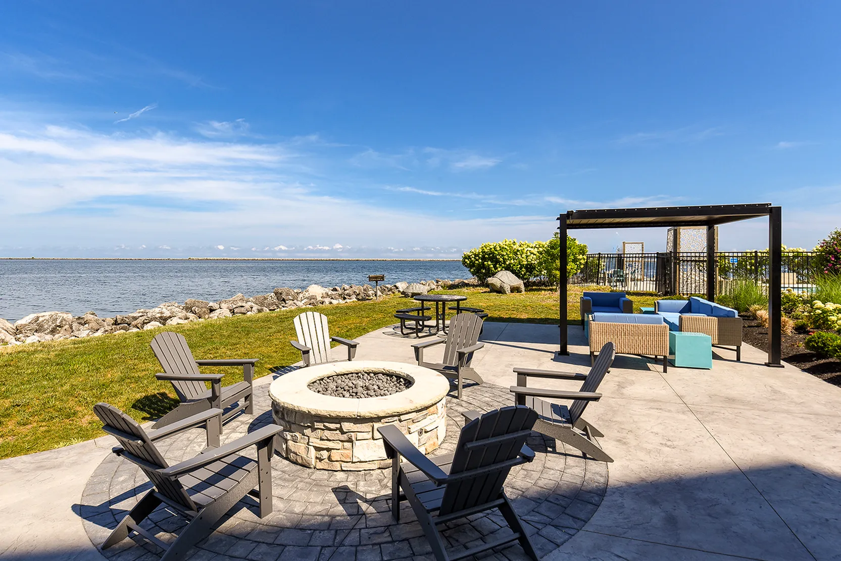 Stunning lakeside patio featuring cozy fire pit surrounded by Adirondack chairs under a clear blue sky, with shaded lounge seating area and vibrant greenery.