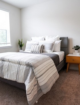Bed with gray and white bedding, gray headboard, wooden nightstand with books and plants, window