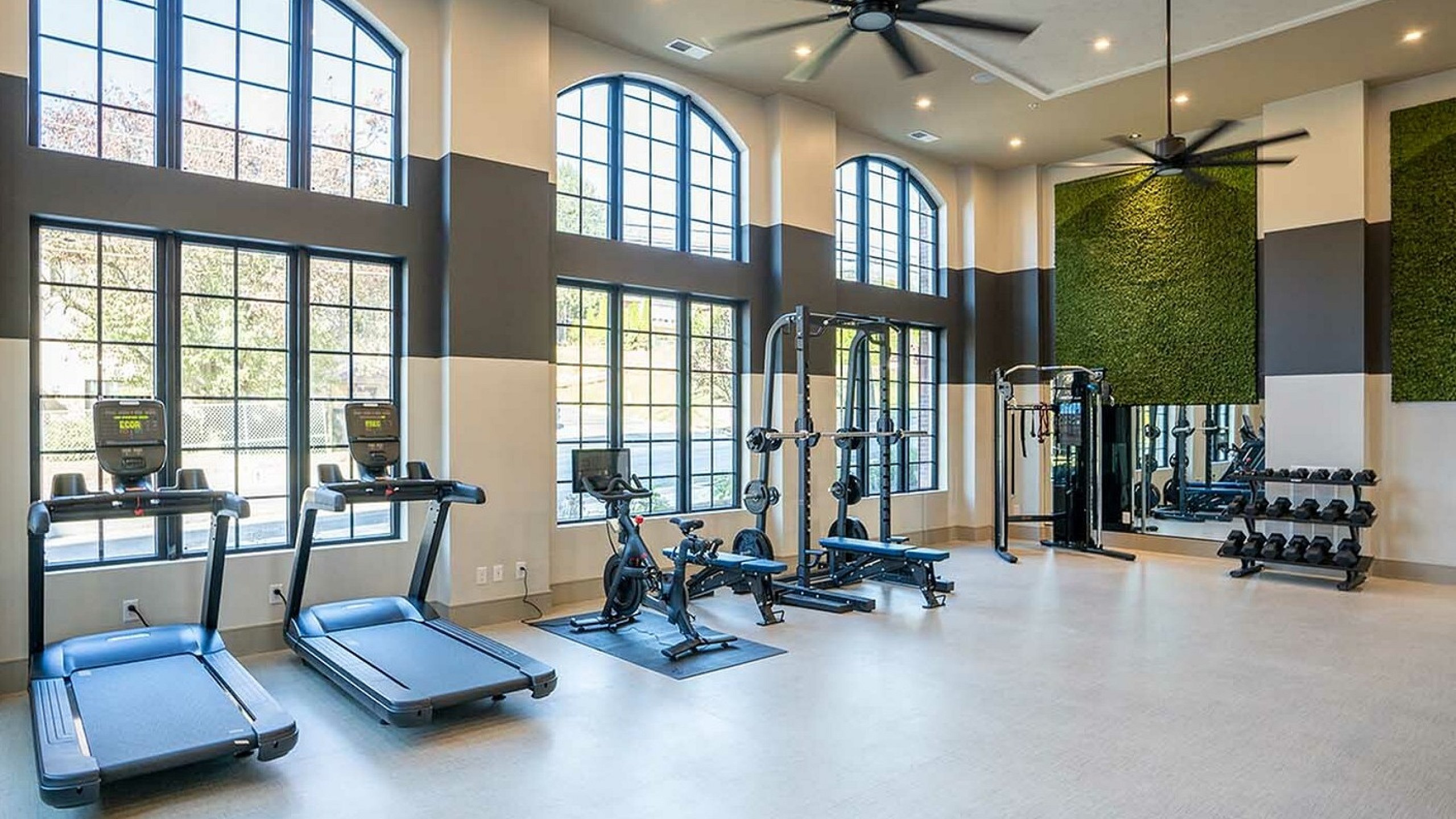 Bright fitness center with large windows, featuring treadmills, exercise bikes, and a weight station.
