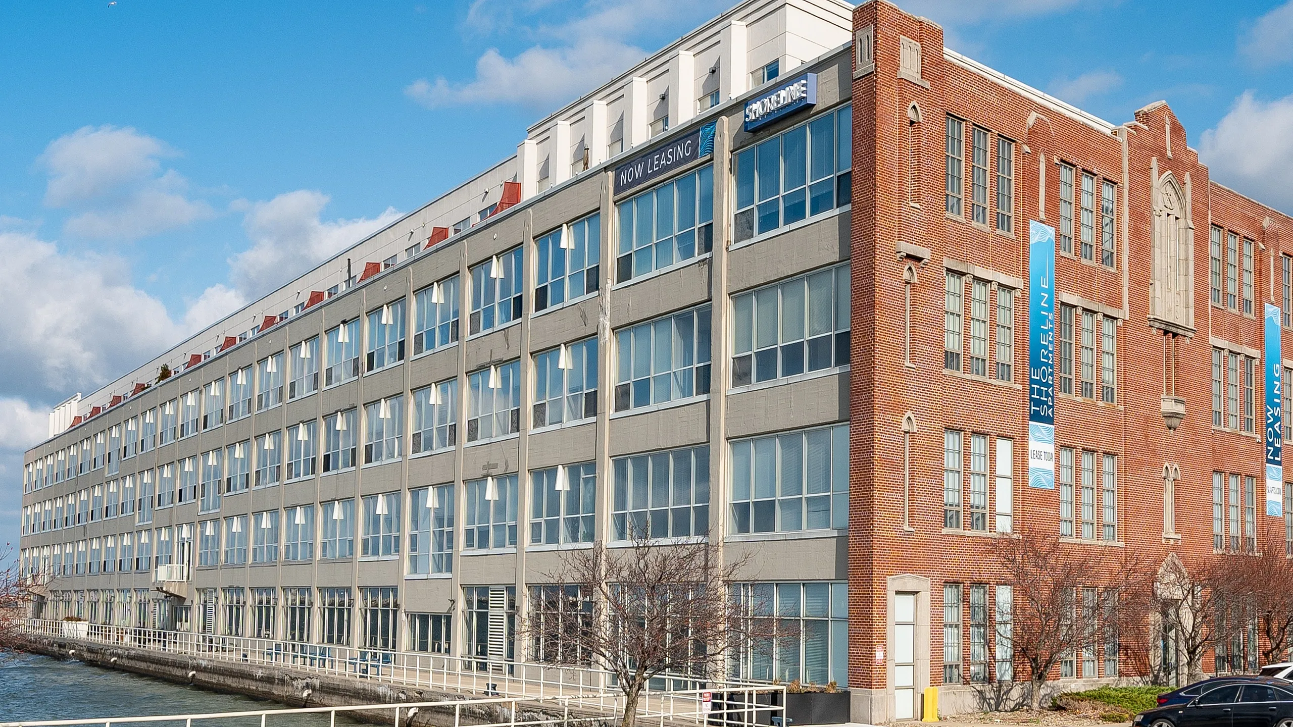 Charming brick and glass apartment building by the waterfront, featuring large windows and vibrant banners announcing leasing opportunities under a clear blue sky.