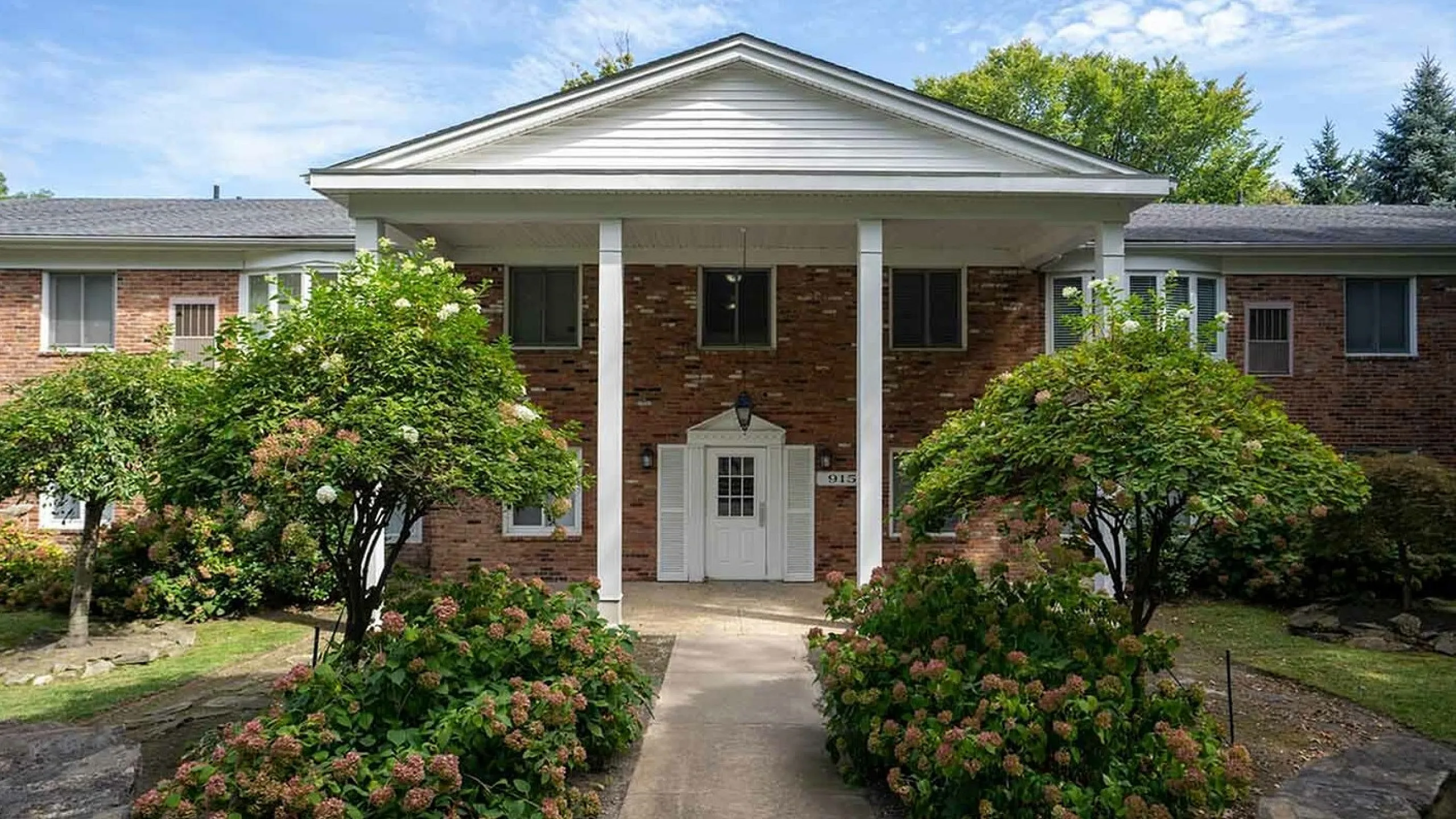 Charming brick apartment building with inviting white columns, surrounded by lush greenery and vibrant flowering shrubs.