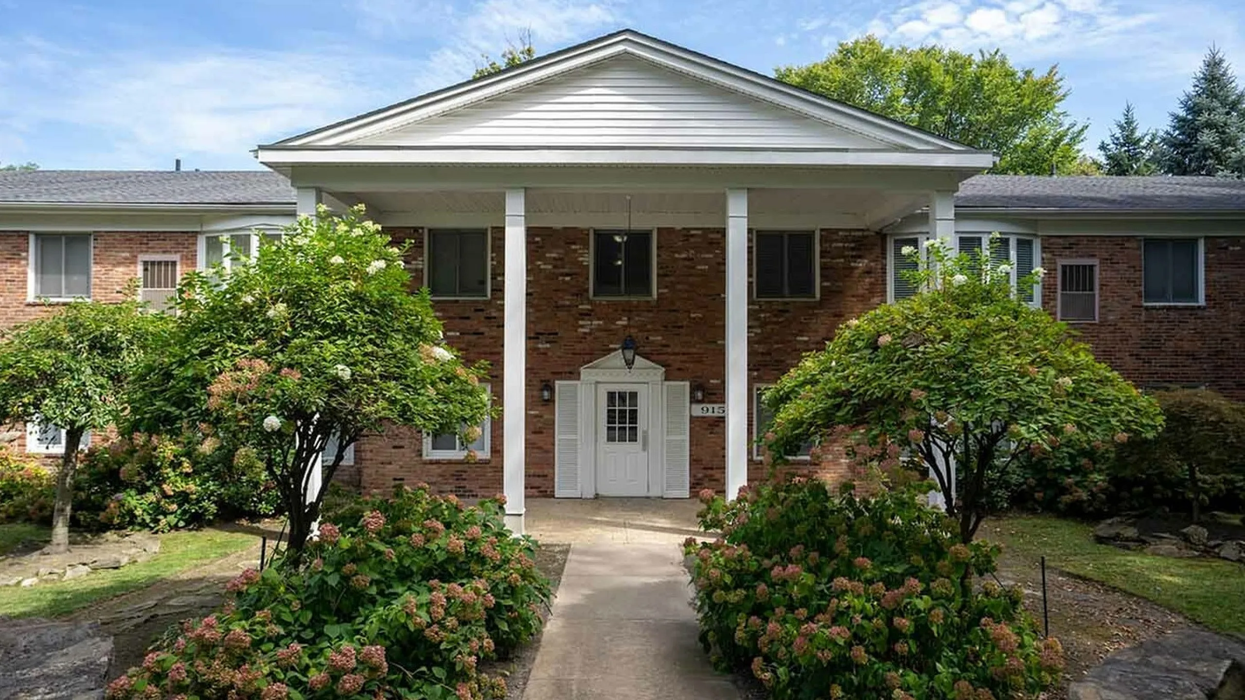 Charming brick apartment building with classic white-pillared entrance, surrounded by lush green landscaping and colorful flowerbeds under a bright blue sky.