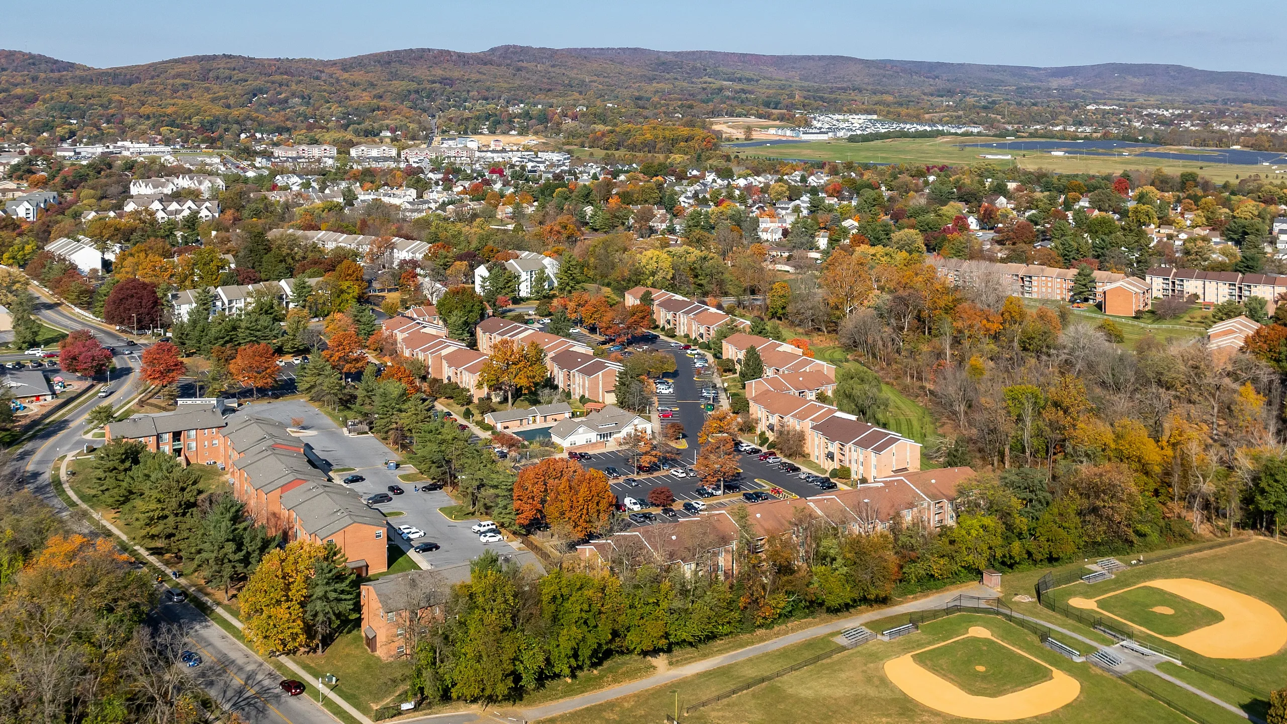 Charming aerial view of a vibrant residential community surrounded by lush greenery and rolling hills, featuring spacious townhomes and well-maintained sports fields.