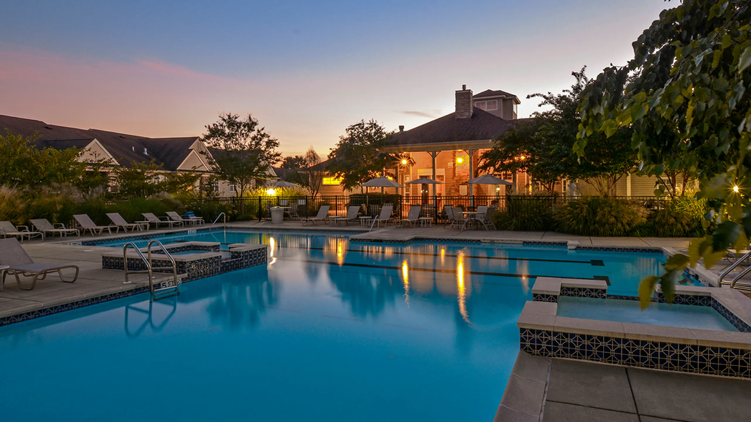 Tranquil evening view of a luxurious apartment complex pool surrounded by lush greenery and stylish loungers.