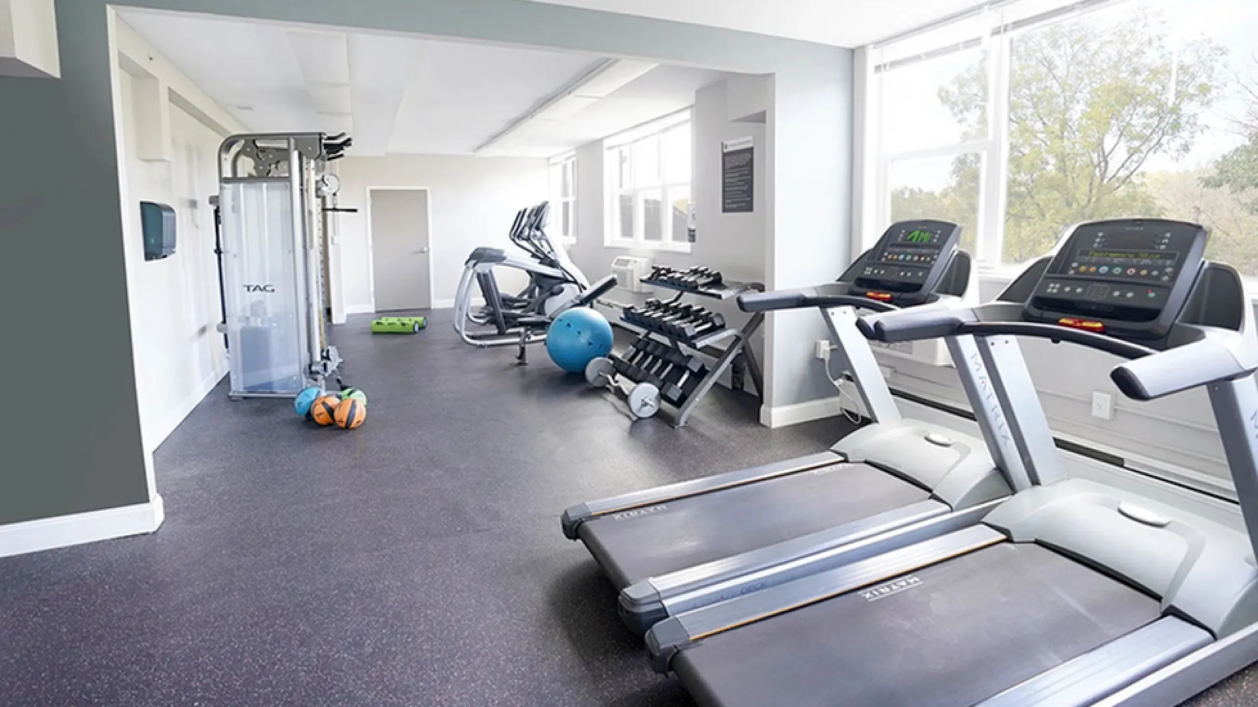 Bright, modern gym featuring treadmills, exercise bikes, and a variety of fitness equipment in a spacious setting with large windows.