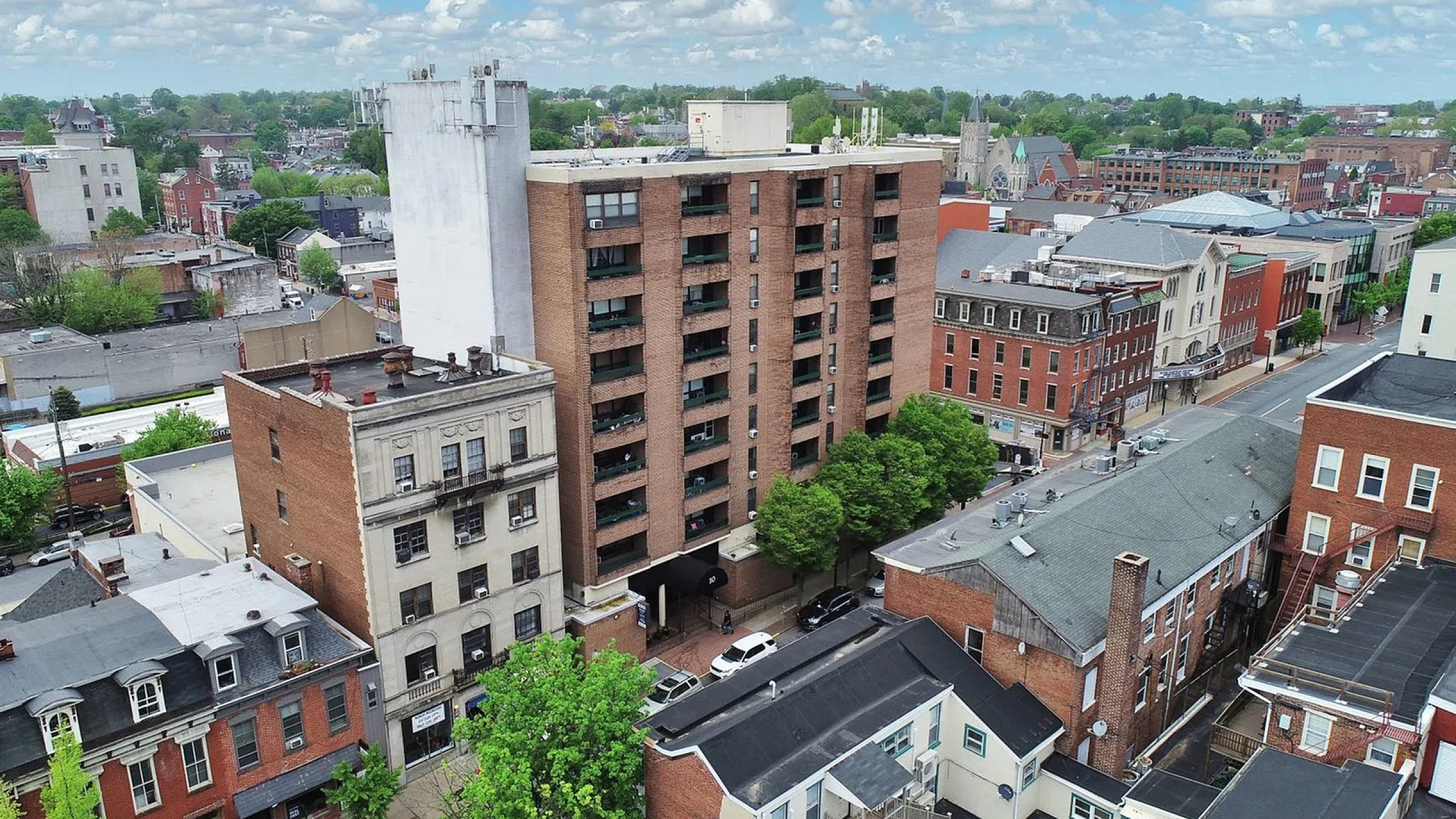 Aerial view of a vibrant urban neighborhood featuring a prominent brick high-rise apartment building surrounded by charming historic architecture and lush greenery.