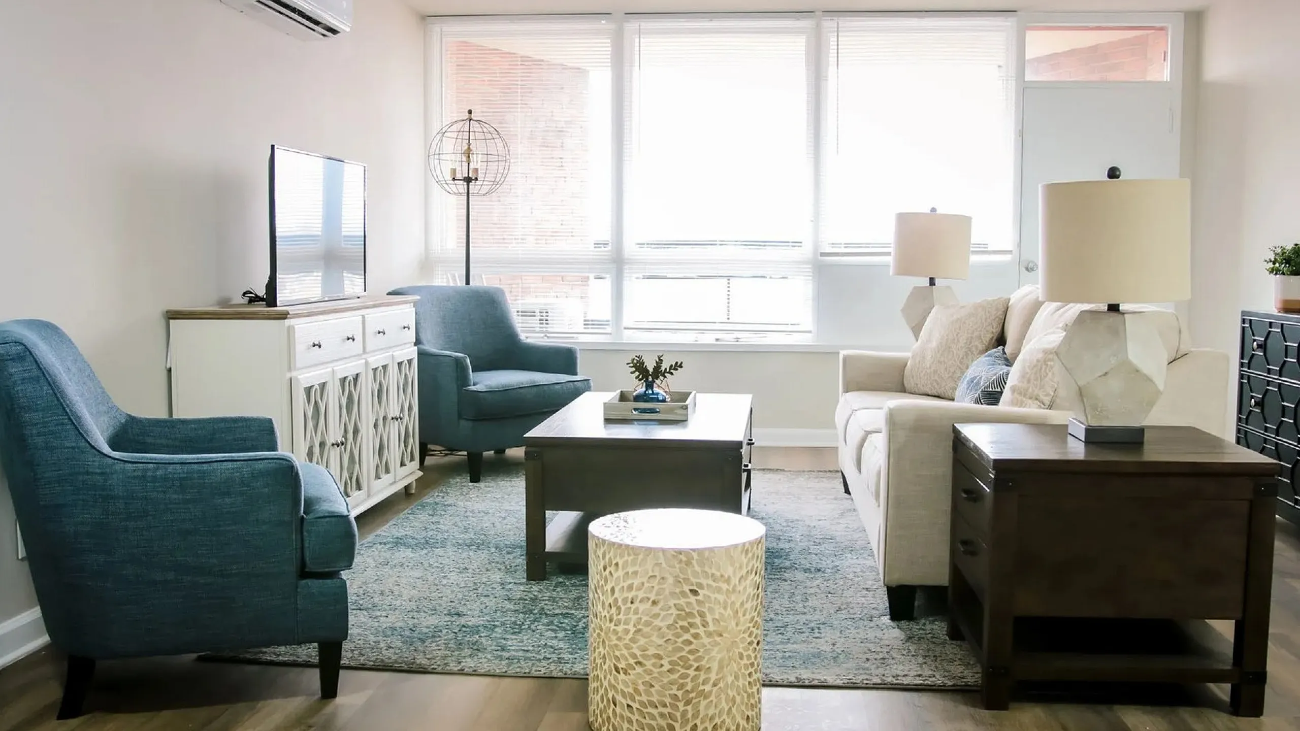 Modern living room with elegant decor featuring blue armchairs, a cozy cream sofa, and natural light from large windows.