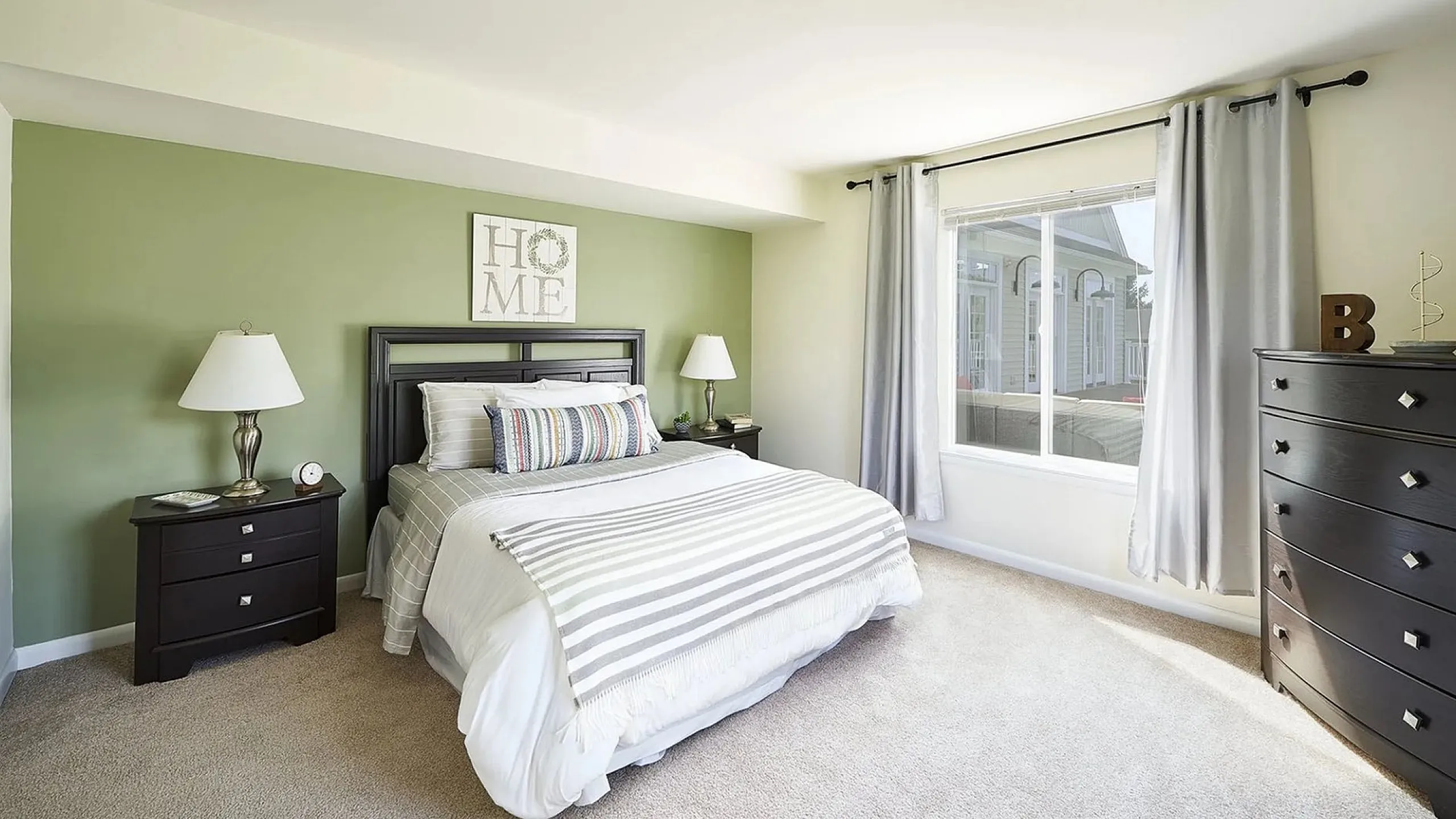Bright, modern bedroom with soft green accent wall, cozy bed, stylish decor, large window, and chic dark wood furniture.