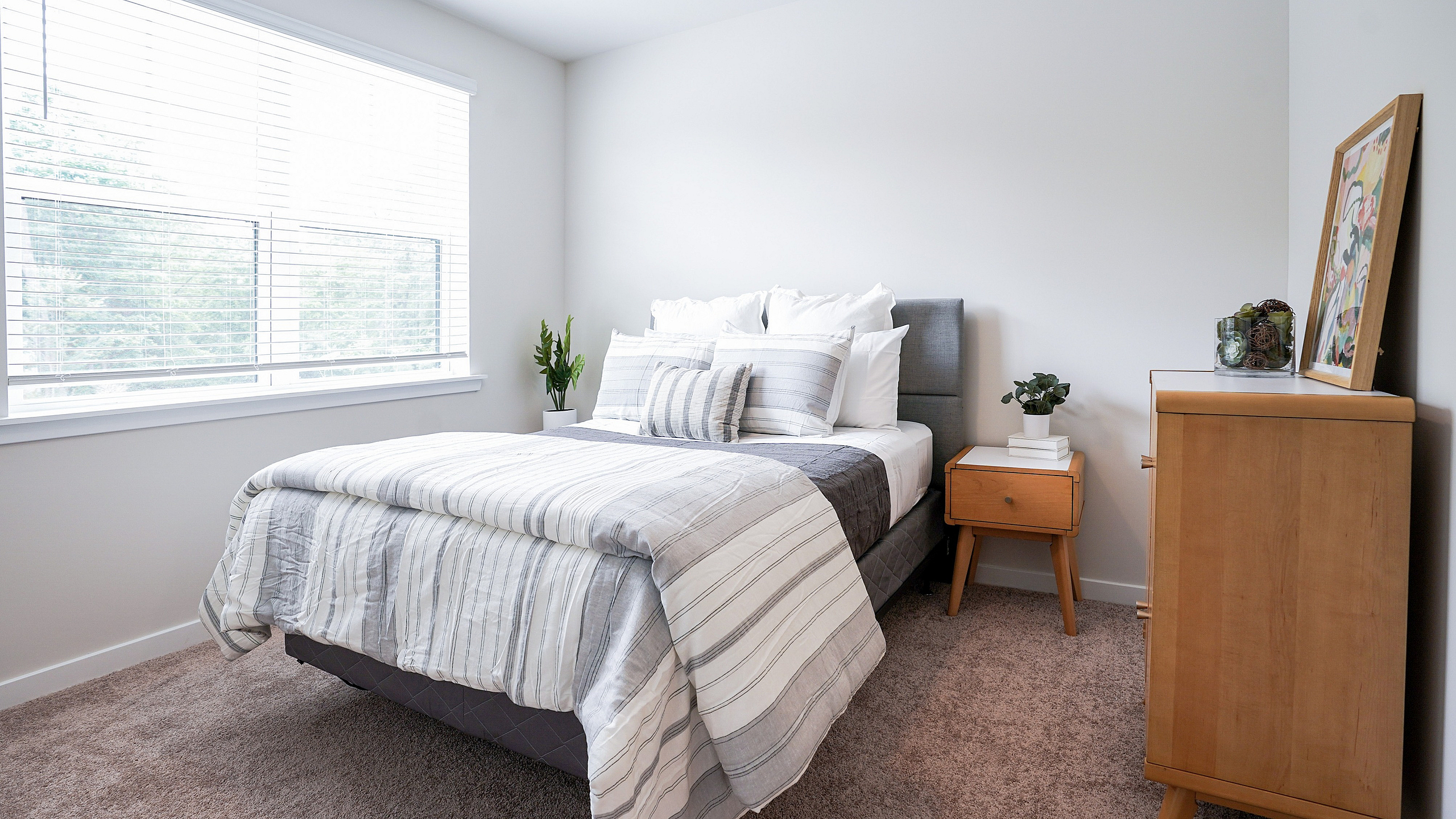 Bright and inviting bedroom featuring a cozy double bed with stylish striped bedding, natural wood furniture, and lush greenery by a large window.