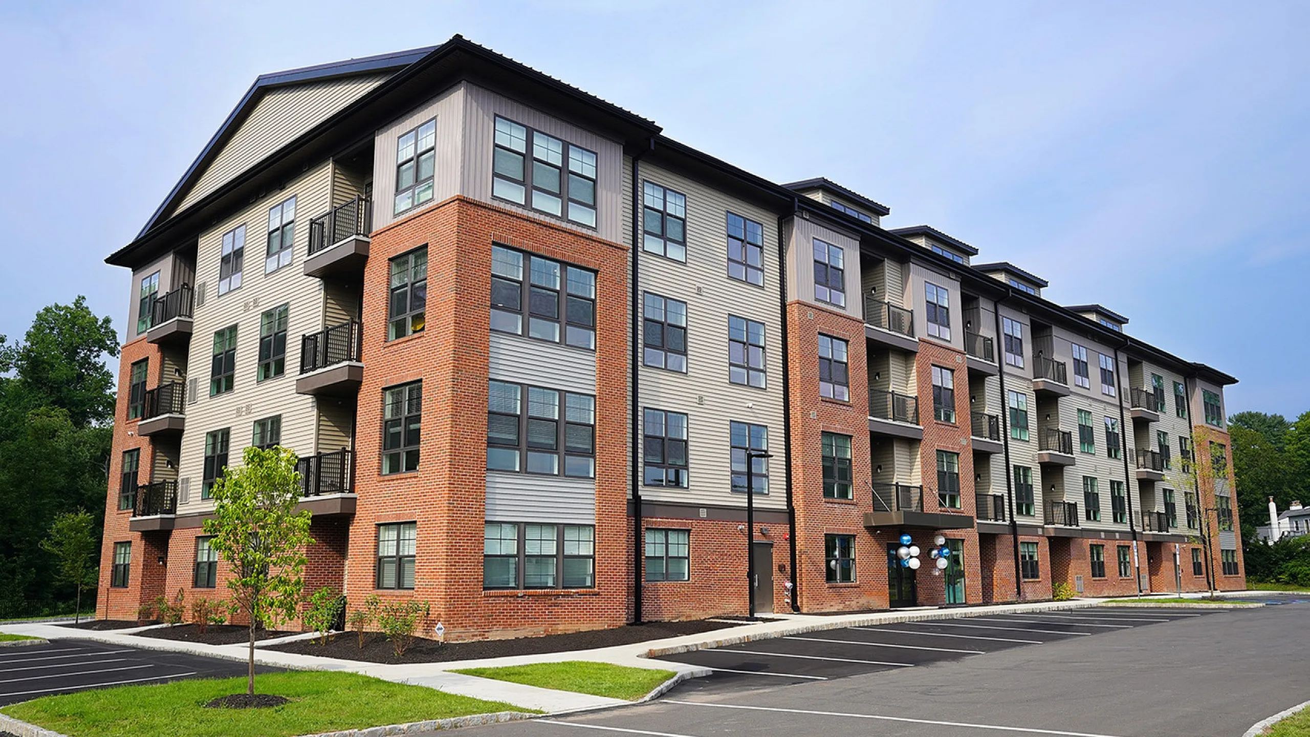 Modern four-story apartment building featuring stylish brick and siding facade with spacious balconies and ample parking, surrounded by lush greenery.