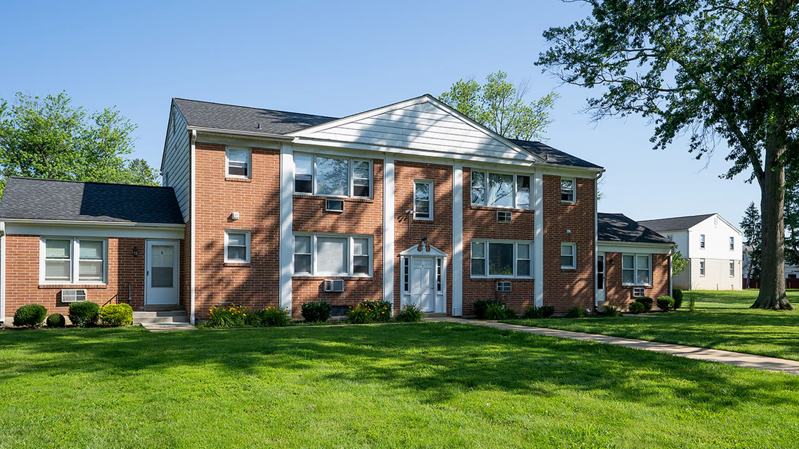 Charming brick apartment building with a classic facade, surrounded by lush greenery and mature trees, offering serene suburban living.