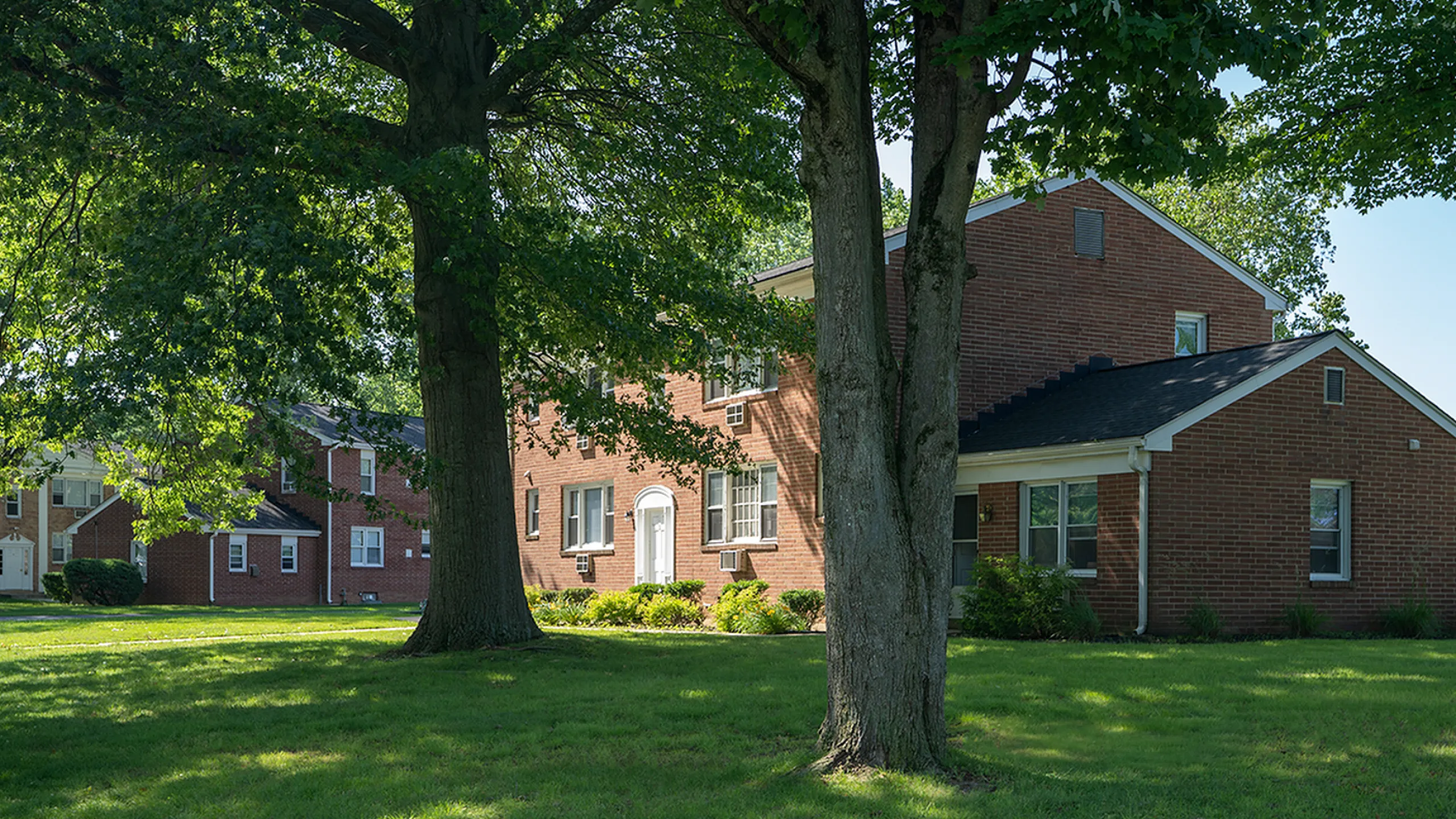 Charming brick apartment complex set amidst lush greenery with mature trees, offering a serene and inviting atmosphere.