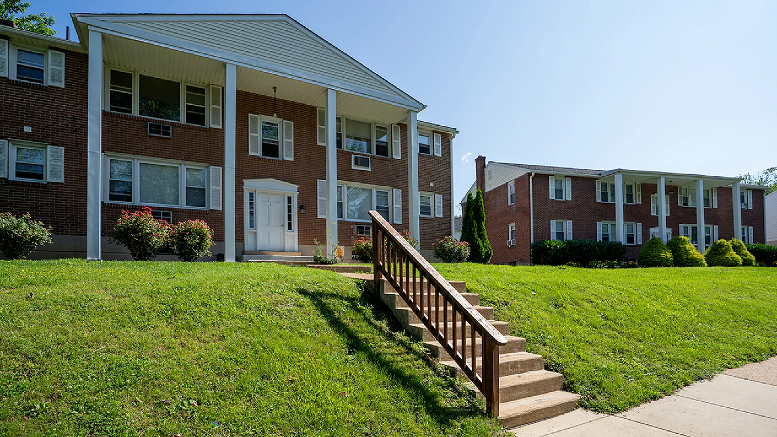 Charming red brick apartment buildings with classic white pillars and lush green lawns under a clear blue sky.