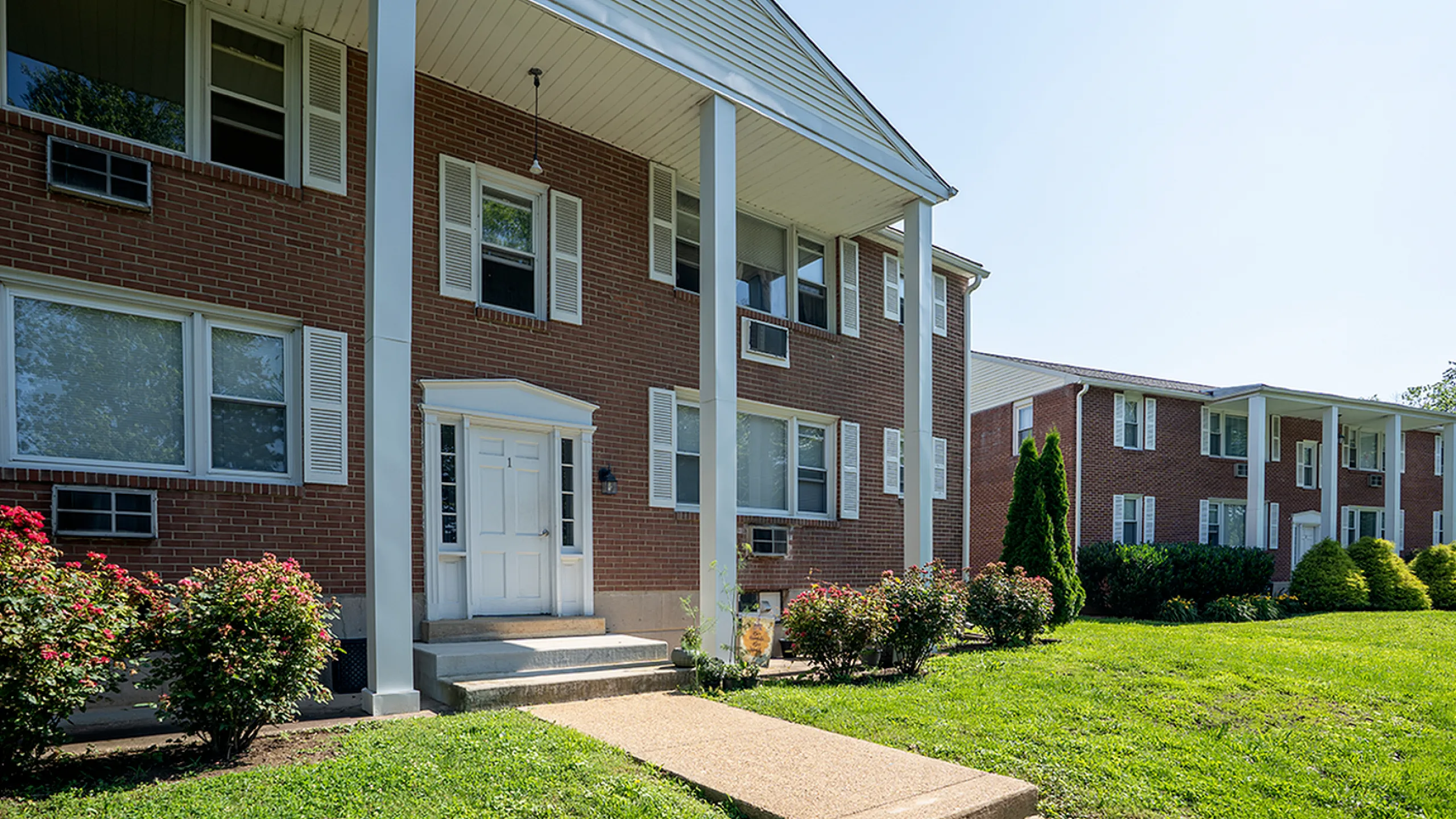 Charming brick apartment building with white accents surrounded by lush greenery and vibrant flower bushes.