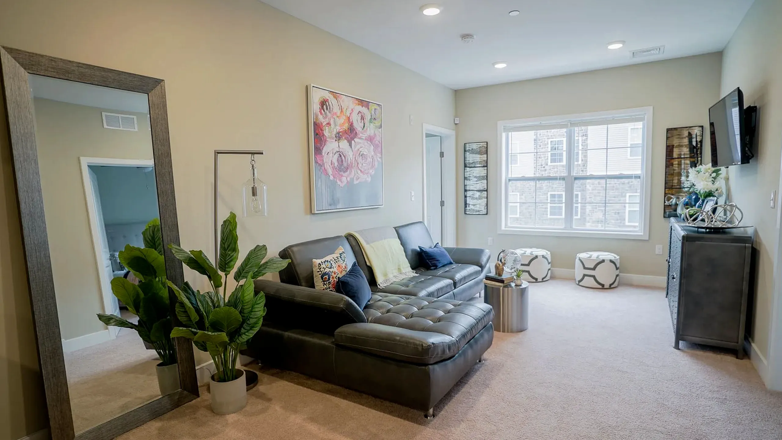 Inviting modern living room with a sleek grey sofa, vibrant artwork, large mirror, and lush indoor plant, flooded with natural light from spacious windows.