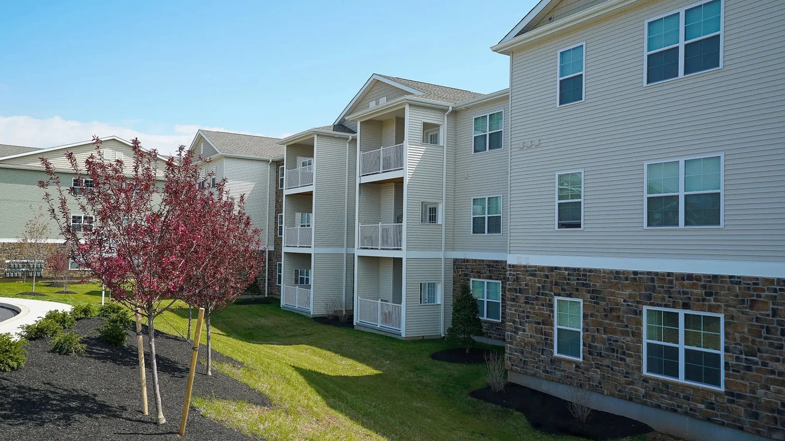 Charming apartment complex with elegant balconies, lush landscaped lawns, and blooming trees under a clear blue sky.