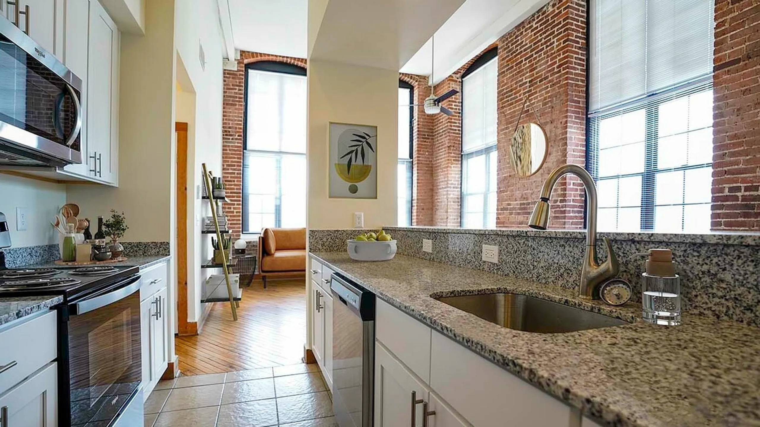 Charming loft-style kitchen with granite countertops, stainless steel appliances, and exposed brick walls leading to a sunlit living area.