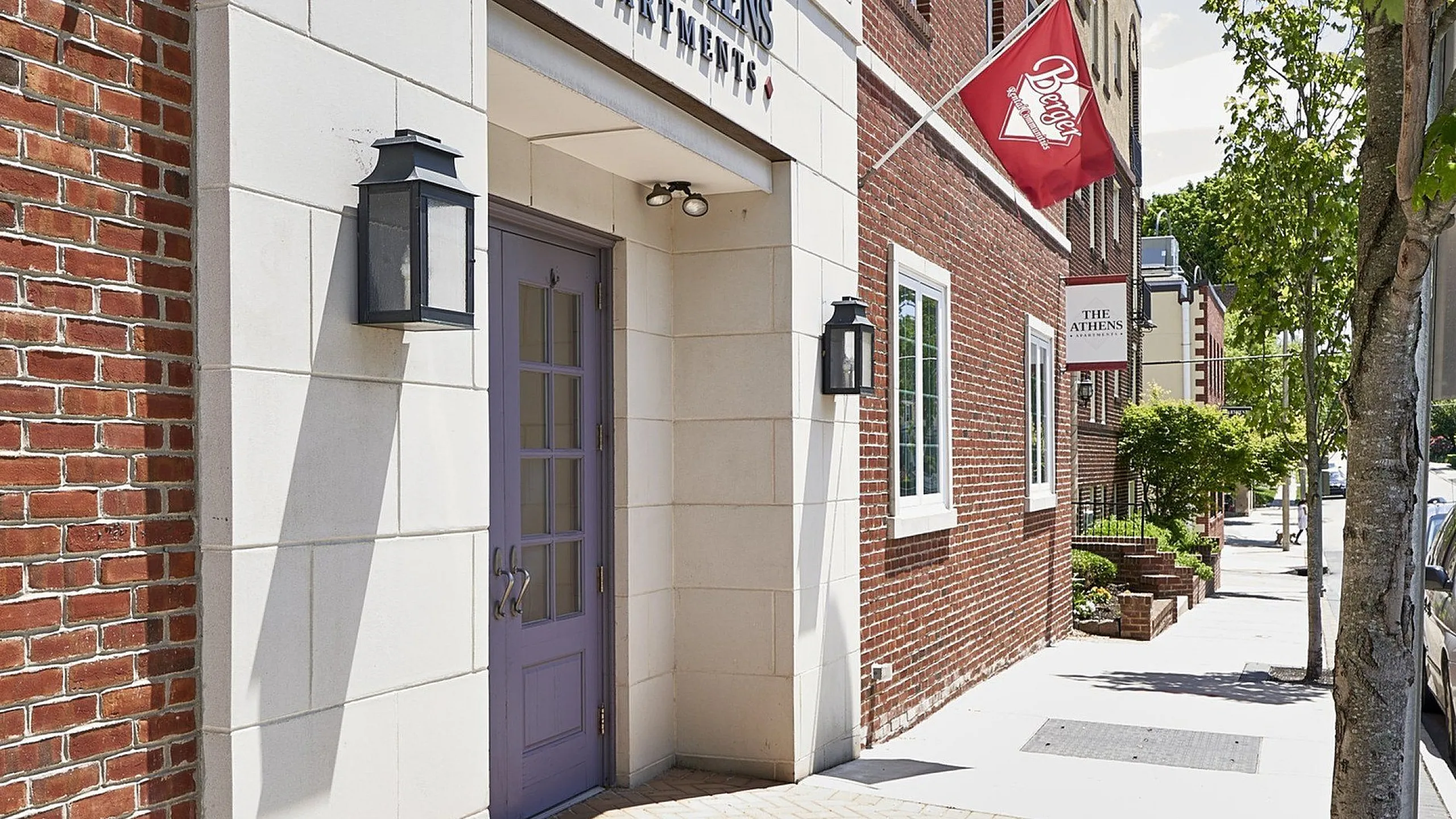 Inviting entrance of The Athens Apartments with charming brick facade and welcoming red flag, surrounded by lush greenery.
