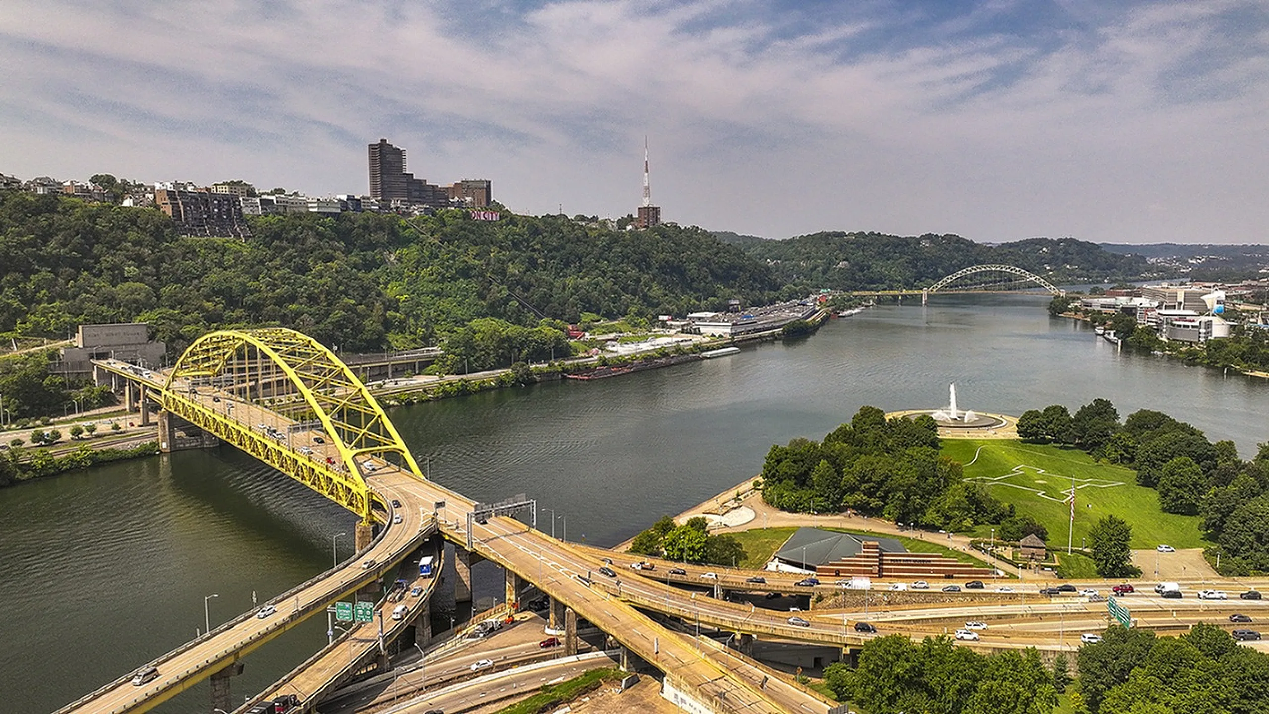 Breathtaking aerial view of a vibrant cityscape featuring a striking yellow bridge spanning a serene river, surrounded by lush greenery and urban architecture.