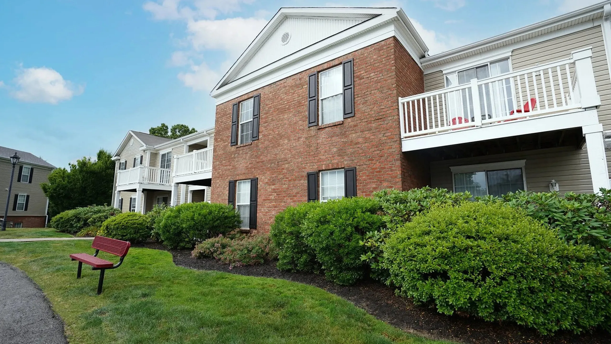 Charming brick apartment building with elegant black shutters, pristine white railings, and lush green landscaping under a bright blue sky.