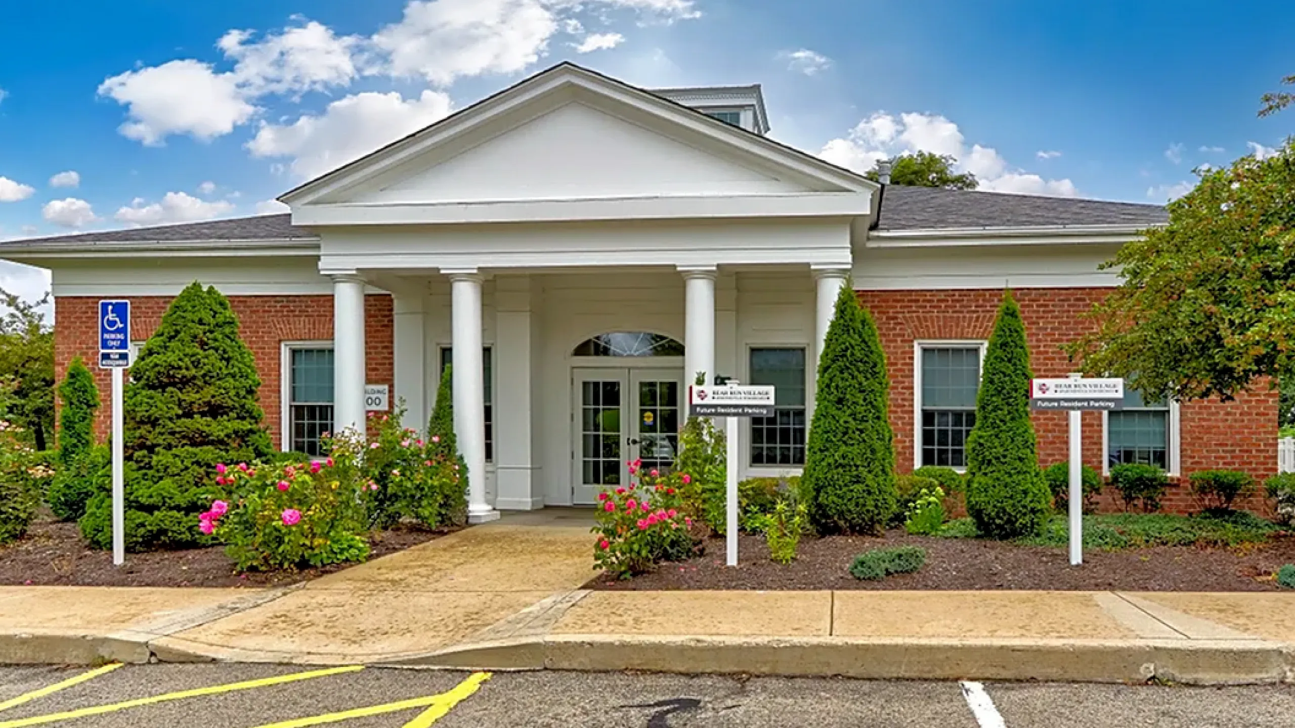 Inviting entrance of a charming brick apartment building with white columns, beautiful landscaping, and clear blue skies.