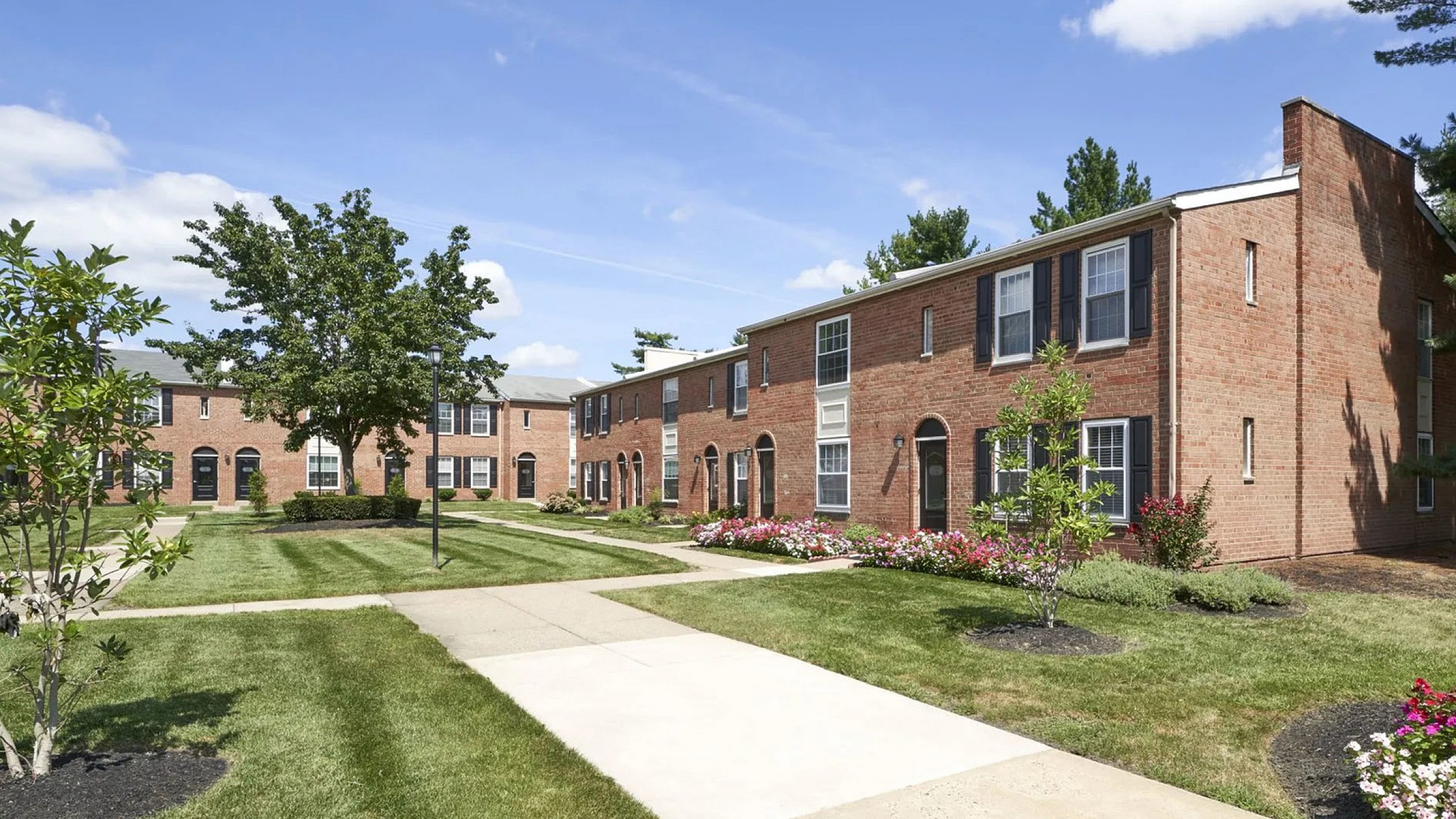 Charming brick apartment complex surrounded by lush green lawns and vibrant flower beds under a clear blue sky.