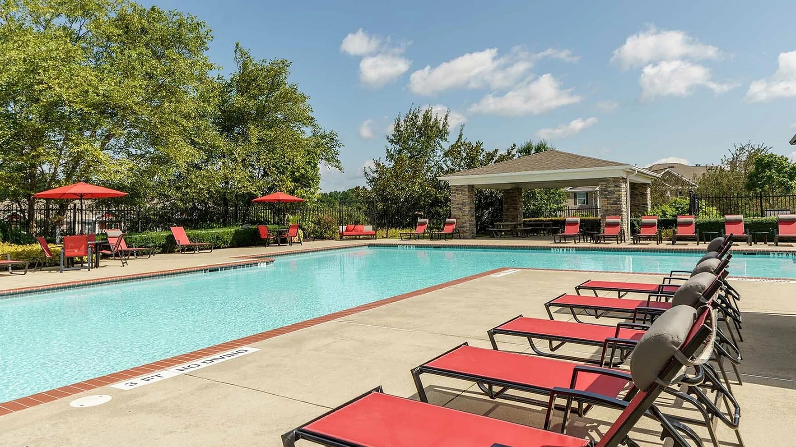 Luxurious apartment community pool area featuring a sparkling blue pool, red sun loungers, and shaded seating amidst lush greenery.