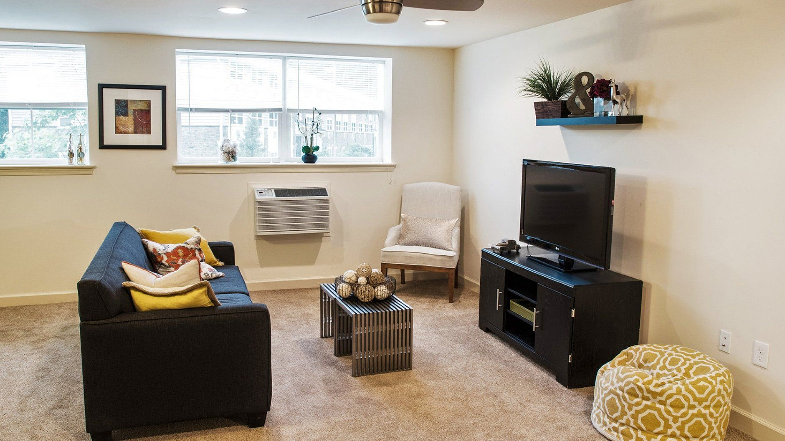 Charming living room with plush carpet, modern ceiling fan, and natural light illuminating cozy seating and a stylish entertainment unit.