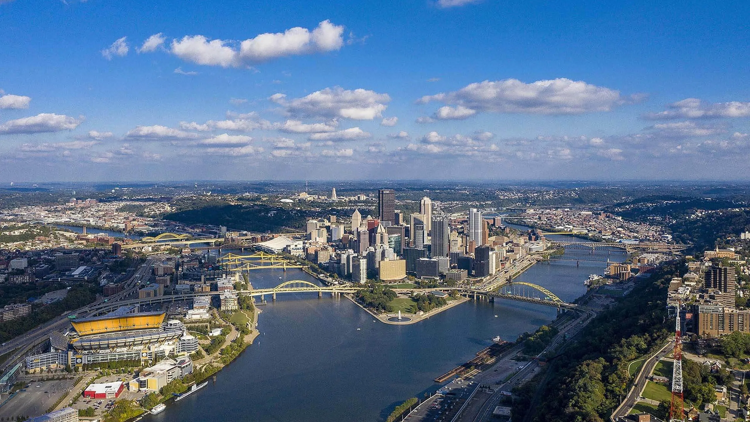 Stunning aerial view of a vibrant cityscape with gleaming skyscrapers, iconic bridges, and a serene river under a bright blue sky, offering a perfect blend of urban living and natural beauty.