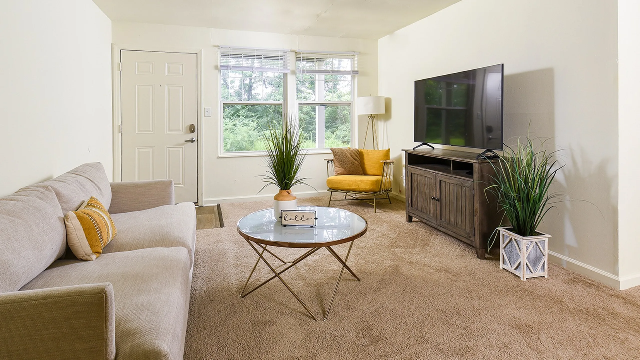 Inviting living room with plush beige sofa, stylish glass coffee table, and modern TV stand. Bright, natural light streams through large windows with serene nature views.