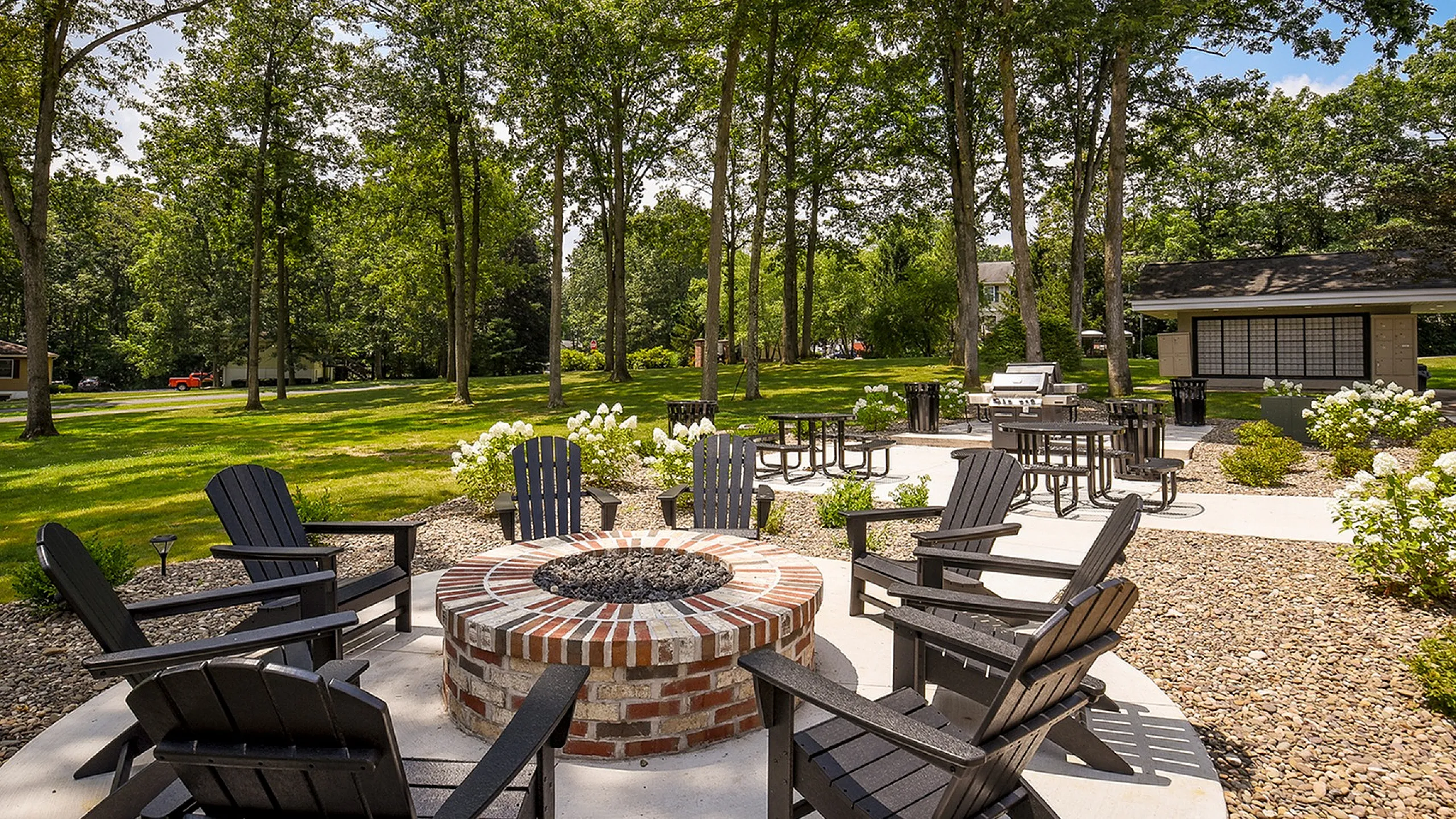 Charming outdoor area with a cozy brick fire pit surrounded by Adirondack chairs, nestled among lush green trees and vibrant landscaping.