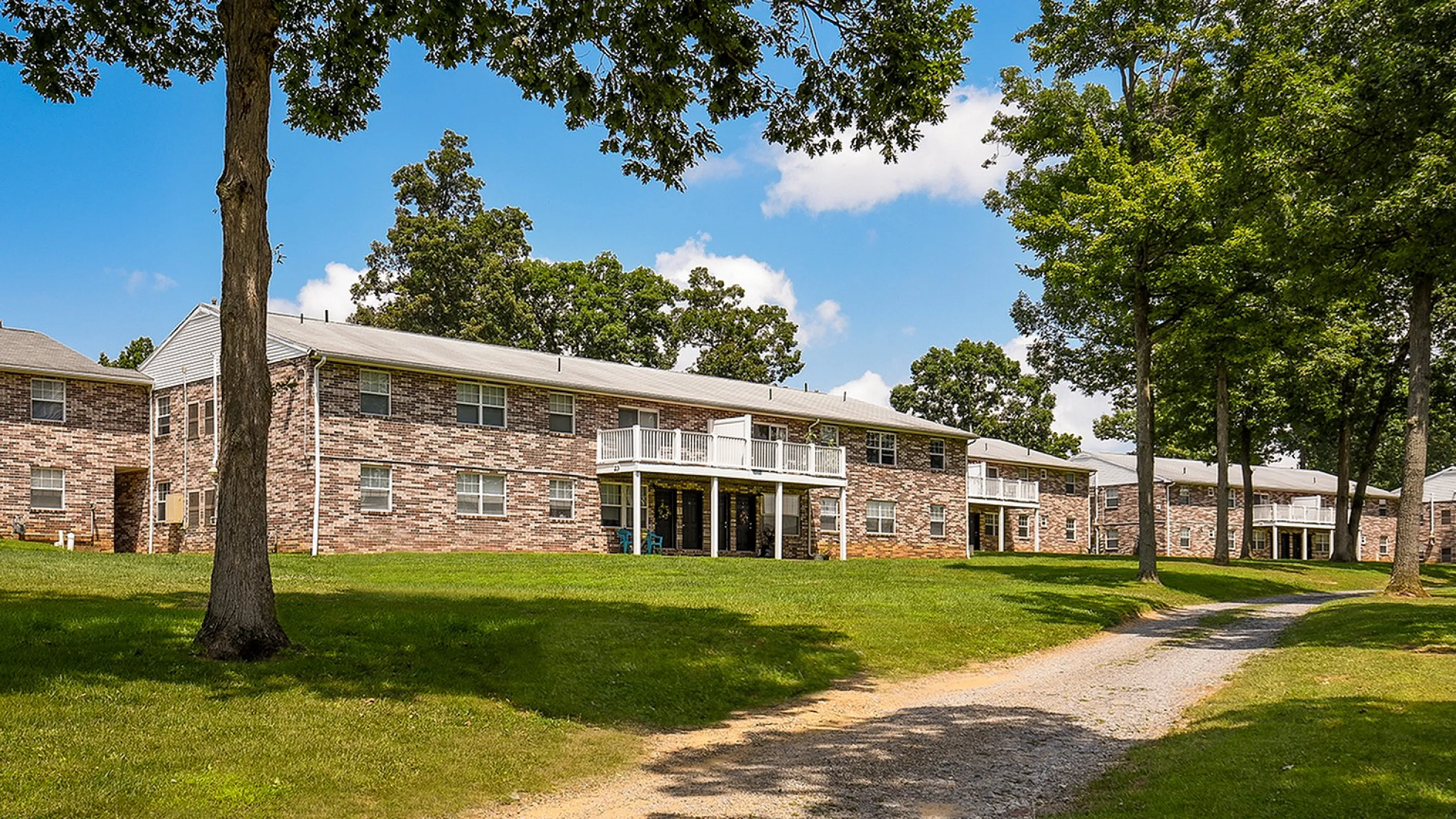 Charming apartment complex with brick exterior surrounded by lush greenery and mature trees, offering a serene and inviting atmosphere.