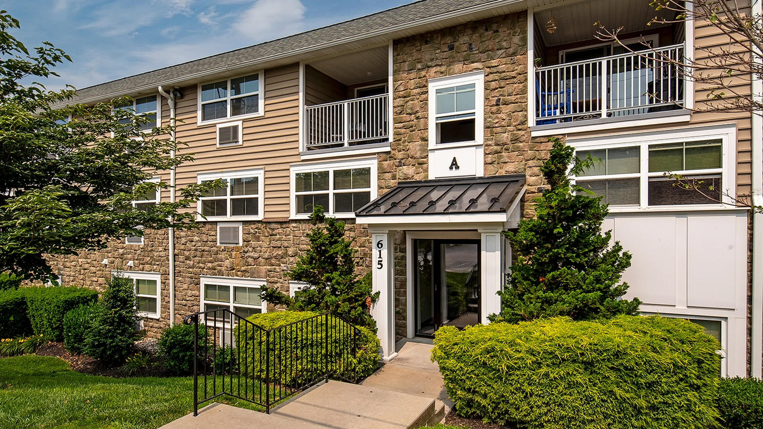 Charming apartment complex exterior featuring stone accents, lush greenery, and inviting entry path.
