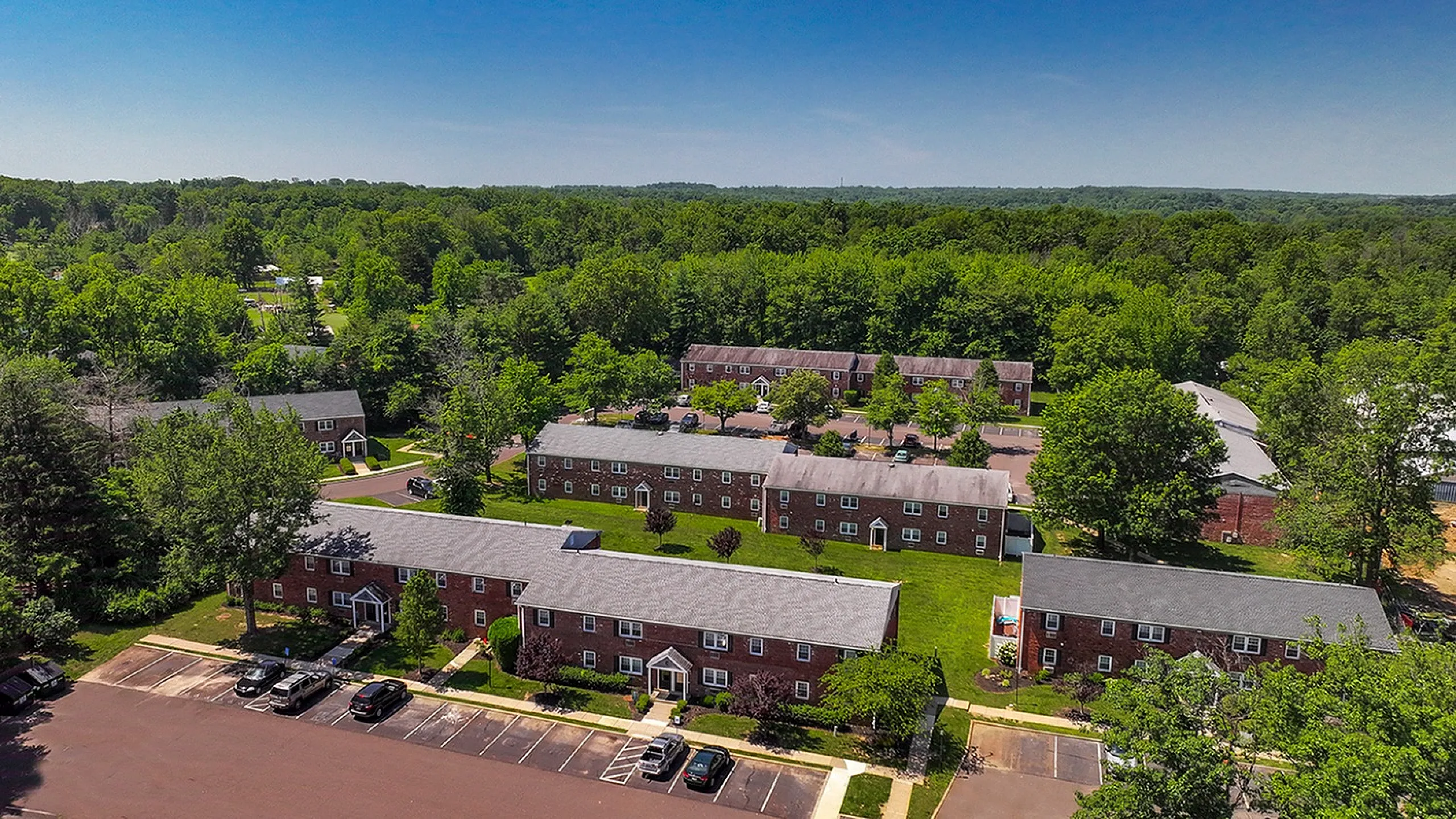 Aerial view of charming brick apartment complex surrounded by lush greenery and ample parking in a serene suburban setting.