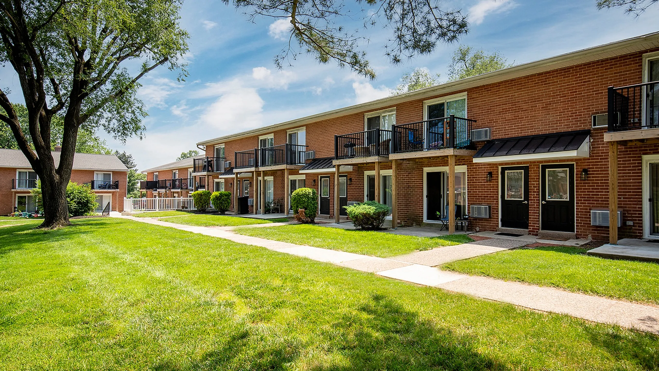 Charming brick apartment complex with private balconies and lush green lawns under a clear blue sky.