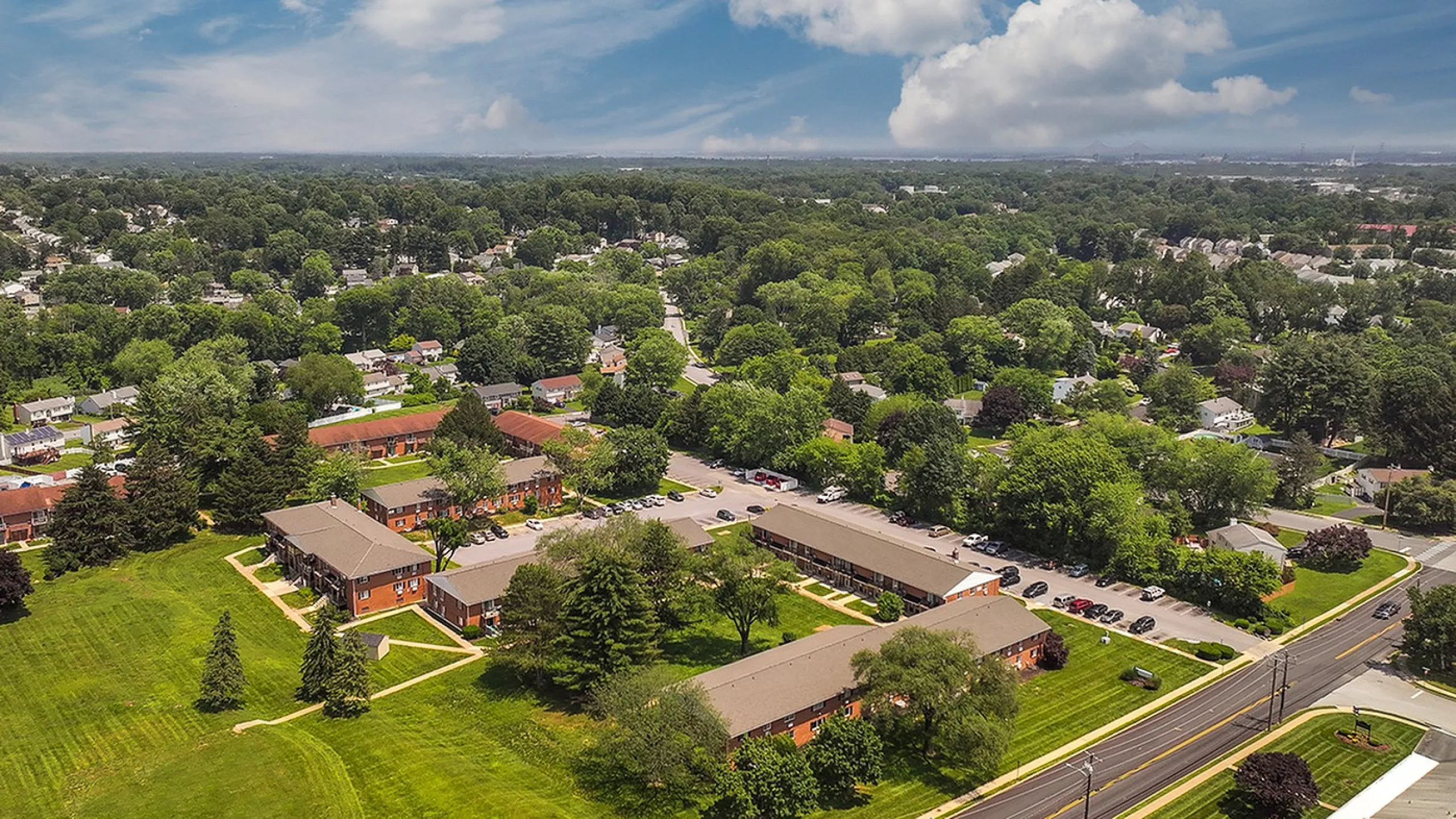 Scenic aerial view of a charming suburban neighborhood with lush green landscapes, modern apartment buildings, and tree-lined streets under a bright blue sky.