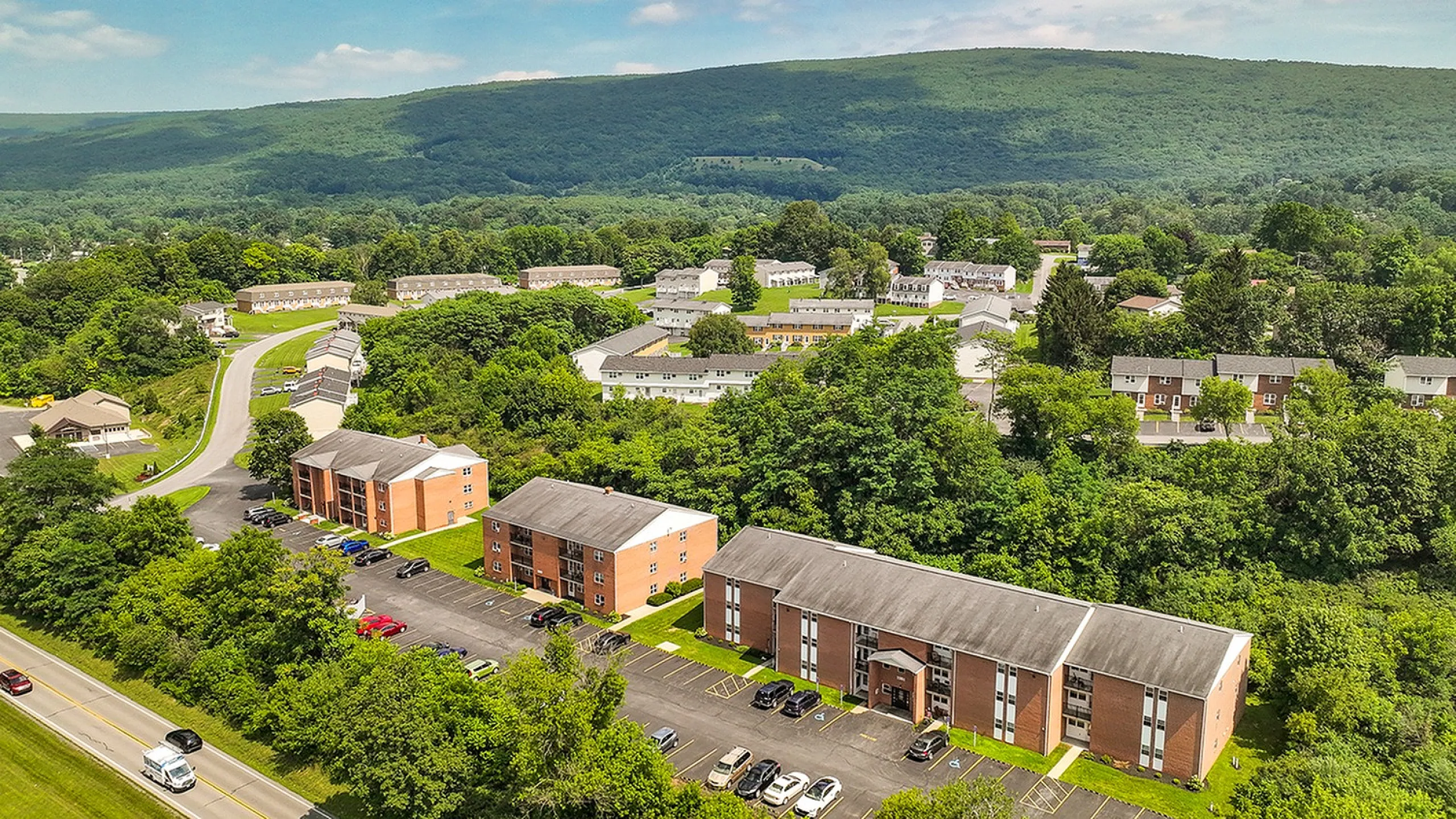 Aerial view of charming apartment complex nestled in lush greenery with scenic mountain backdrop, highlighting tranquil and picturesque living environment.