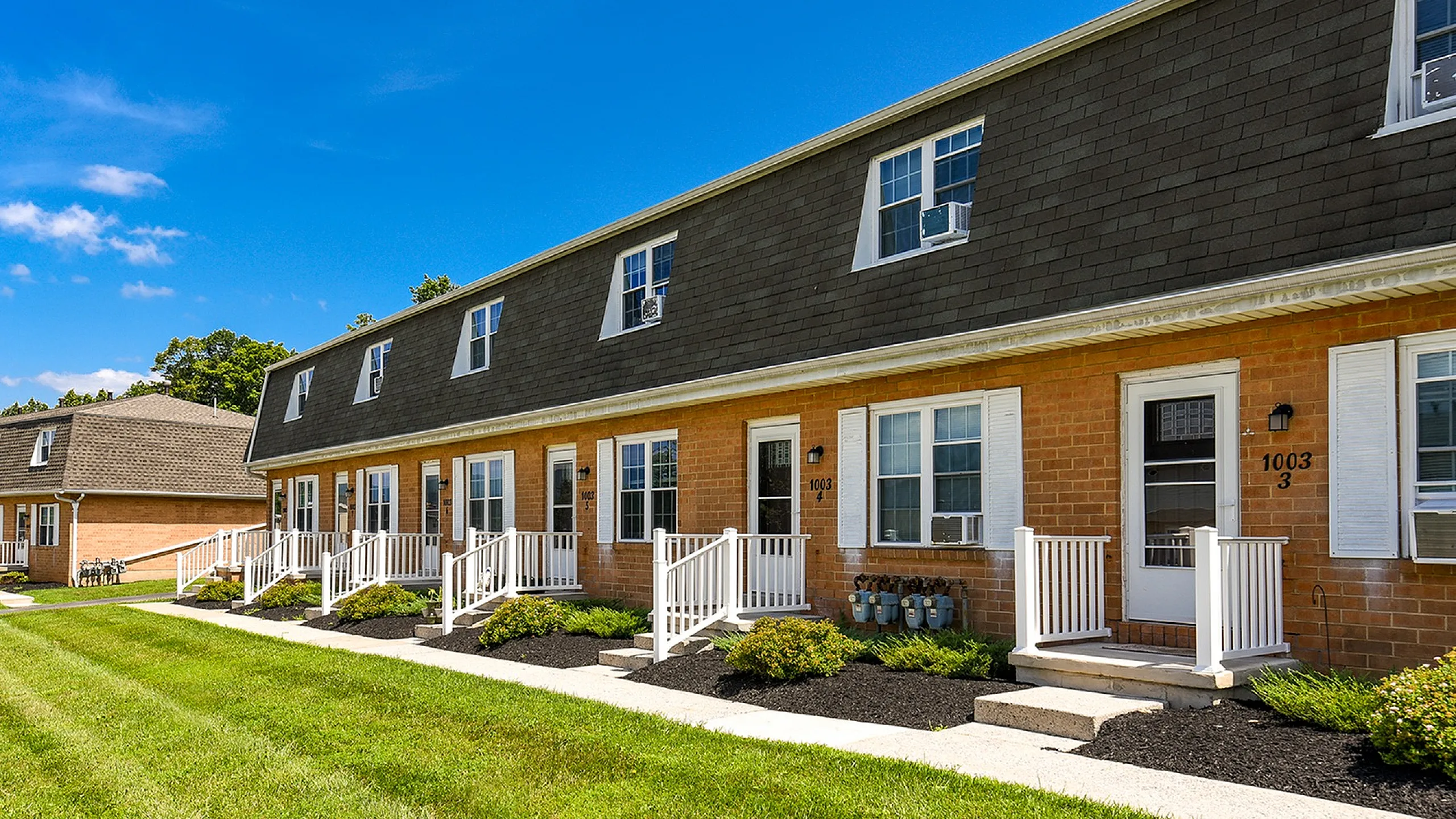 Charming townhouse complex with inviting white porches, surrounded by lush green lawns and a bright blue sky.